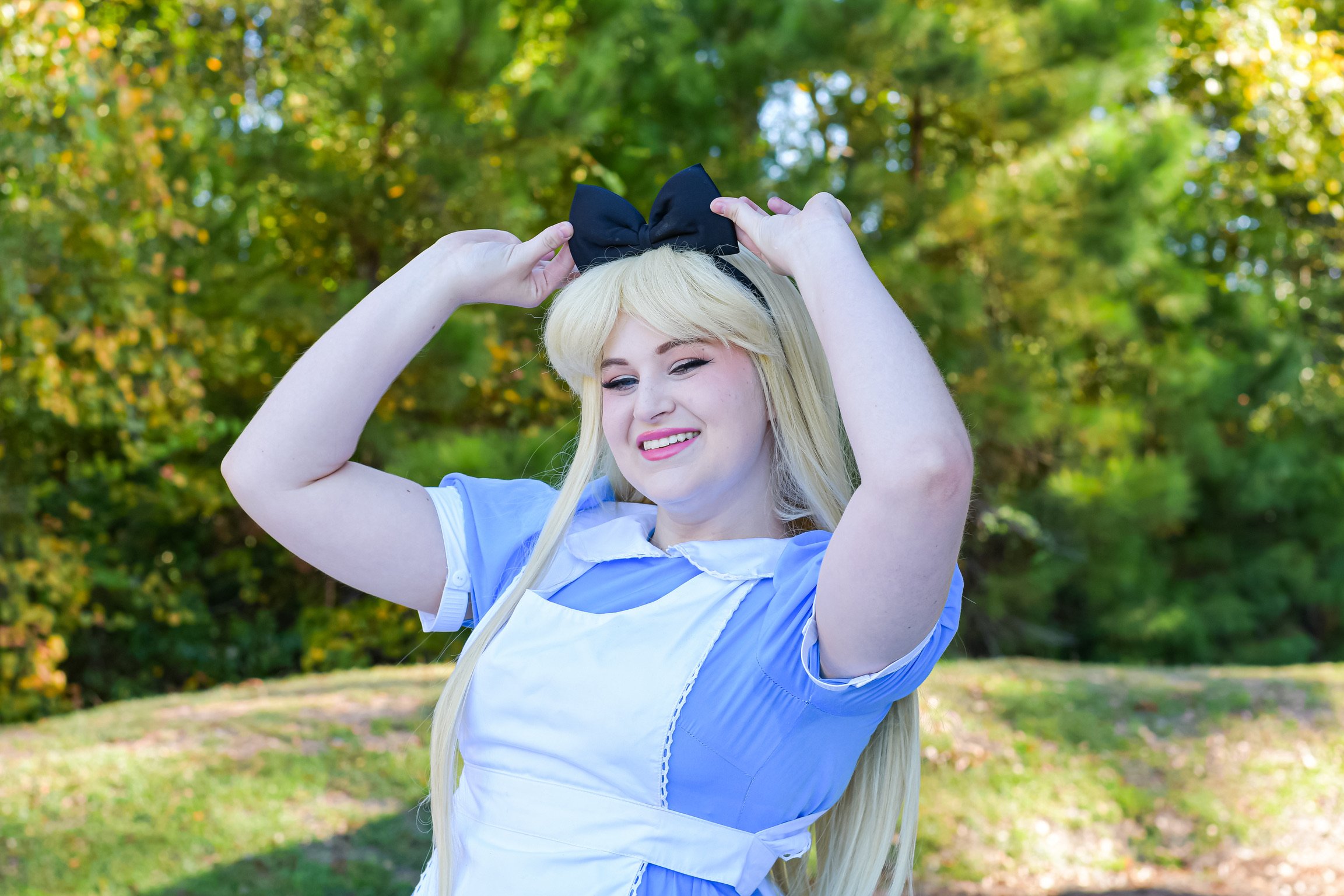 A young woman with long blonde hair dressed in a blue and white costume, adjusting a black bow headband, outdoors in a green park, smiling.