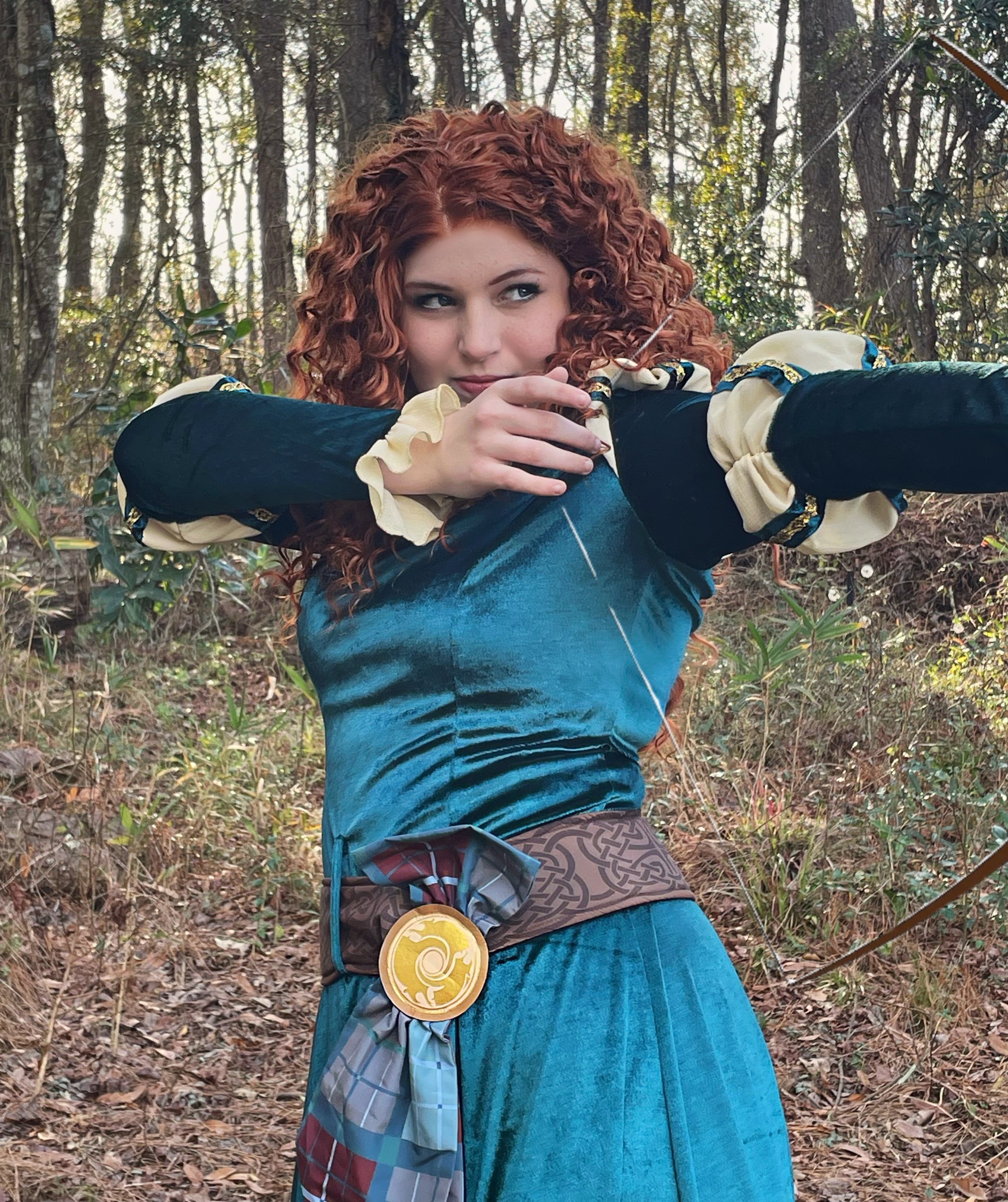 A woman with curly red hair dressed in a medieval-style costume, holding a bow in a forested area.
