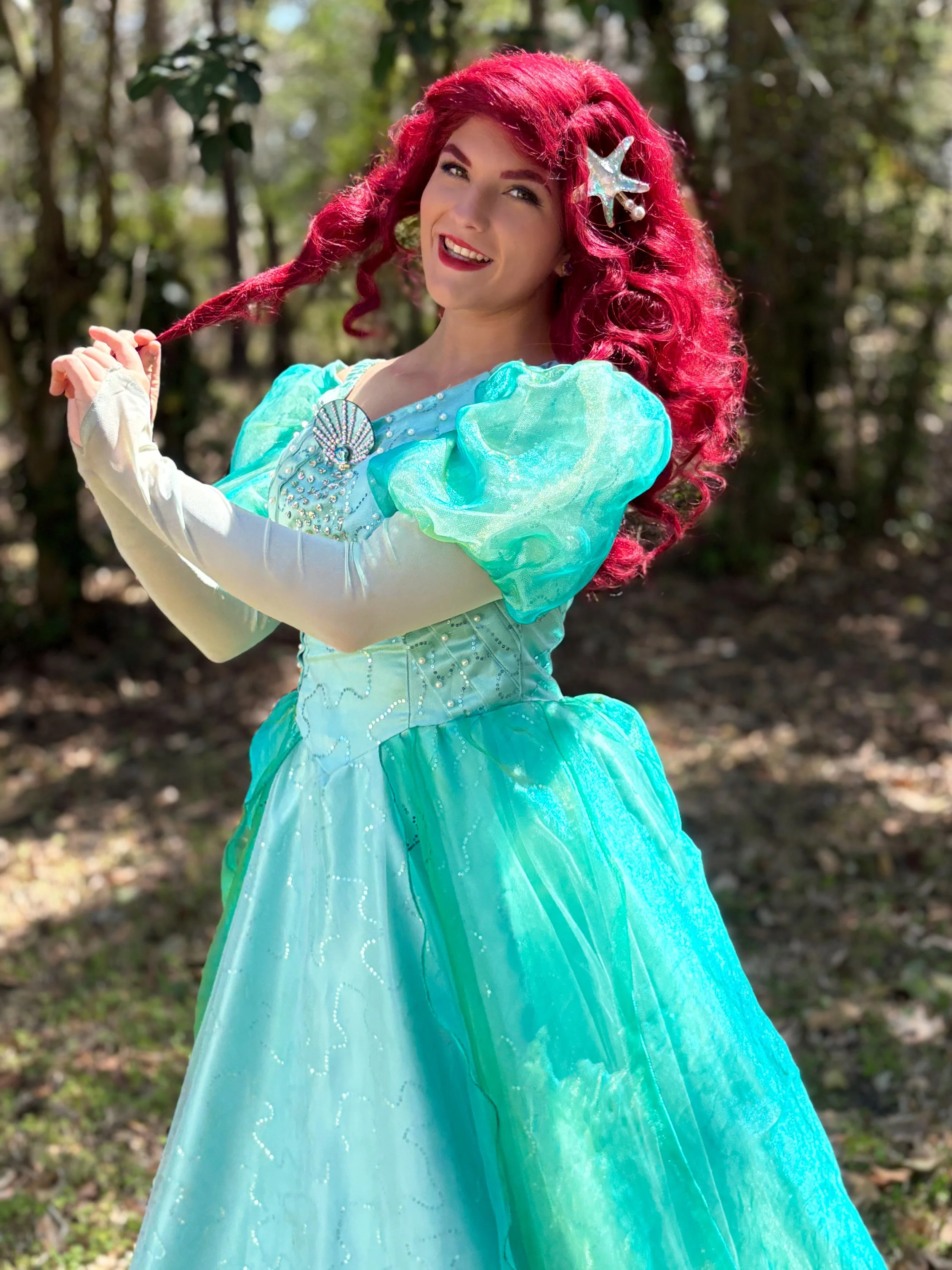 A woman with bright red hair dressed as a fairy or mermaid, wearing a flowing turquoise dress and a starfish hair accessory, standing in a grassy field at sunset with a bokeh effect in the background.