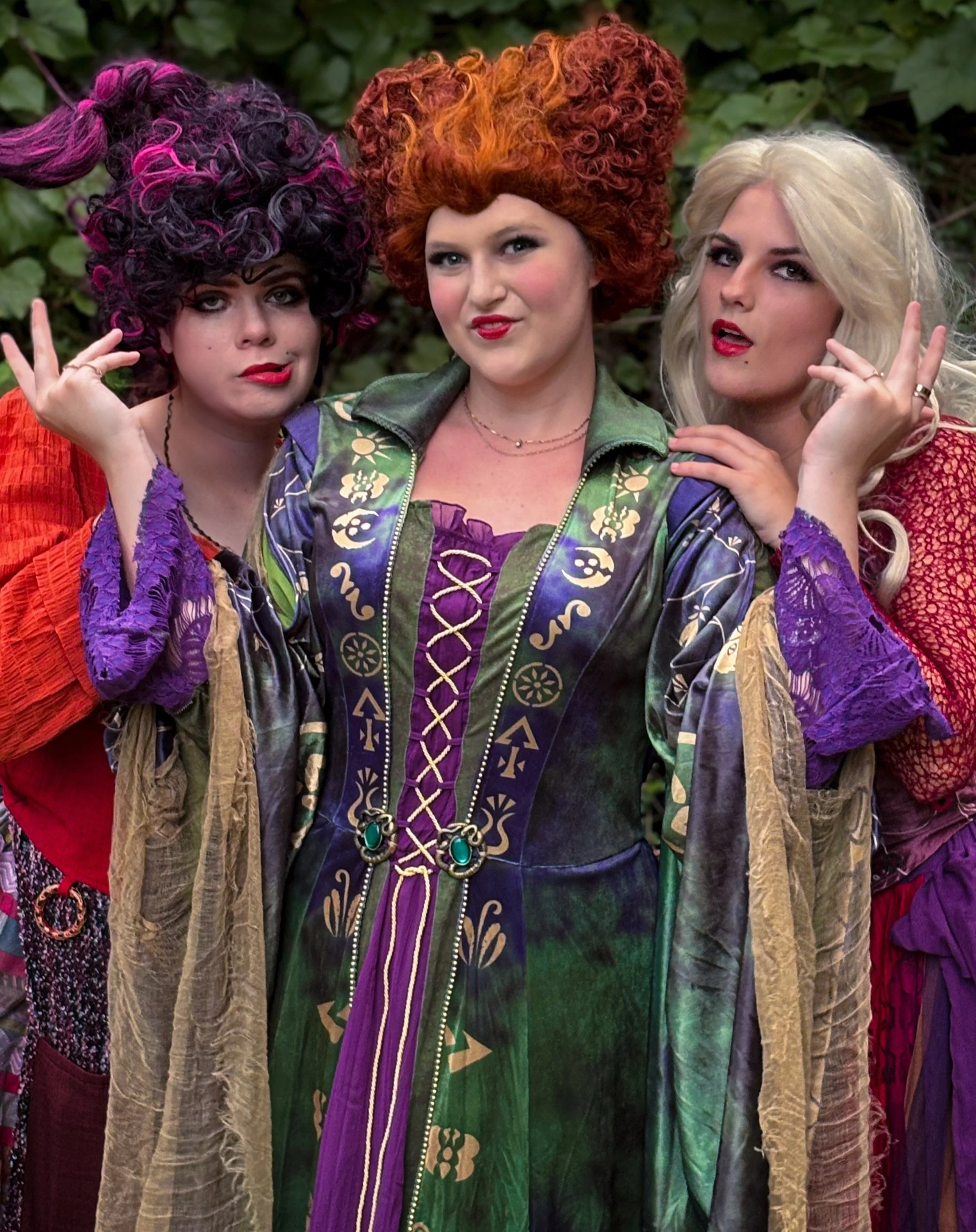 Three women dressed in colorful, extravagant costumes; two of them have curly, brightly colored hair and the third has long, platinum blonde hair. They are posing outdoors with green foliage in the background.