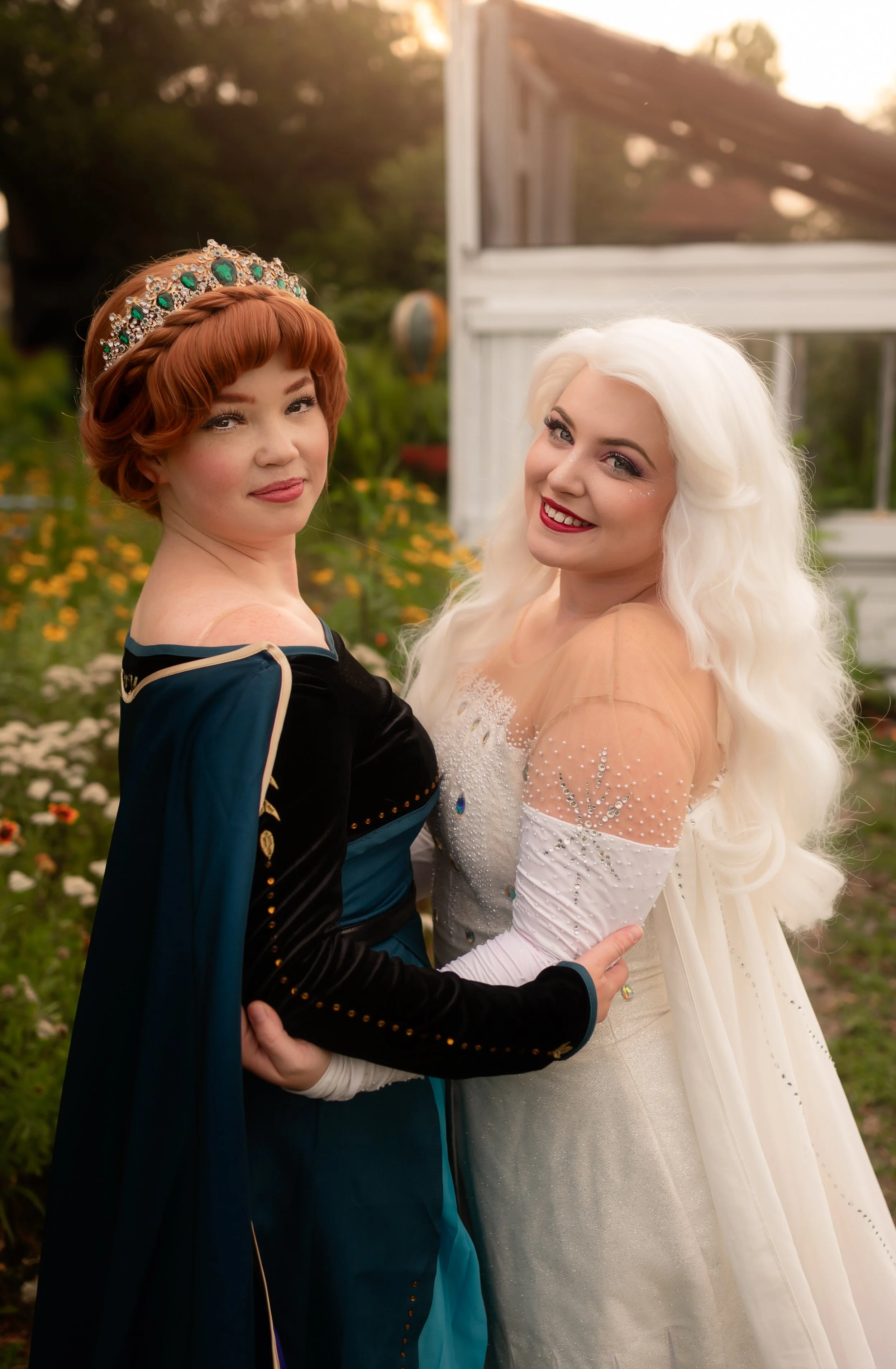 Two women dressed as Disney princesses, one as Anna from Frozen and the other as Elsa, standing outdoors in a garden during sunset.