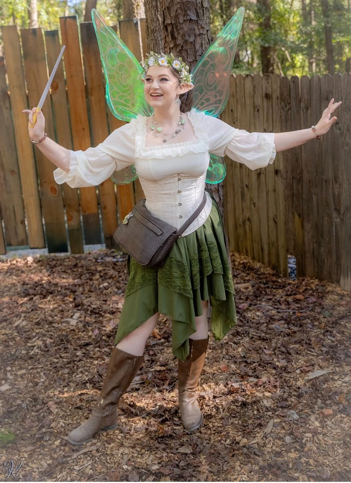 A person dressed as a fairy with iridescent wings, a flower crown, elf ears, and a sword standing outdoors on a bed of leaves, with a wooden fence and trees in the background.