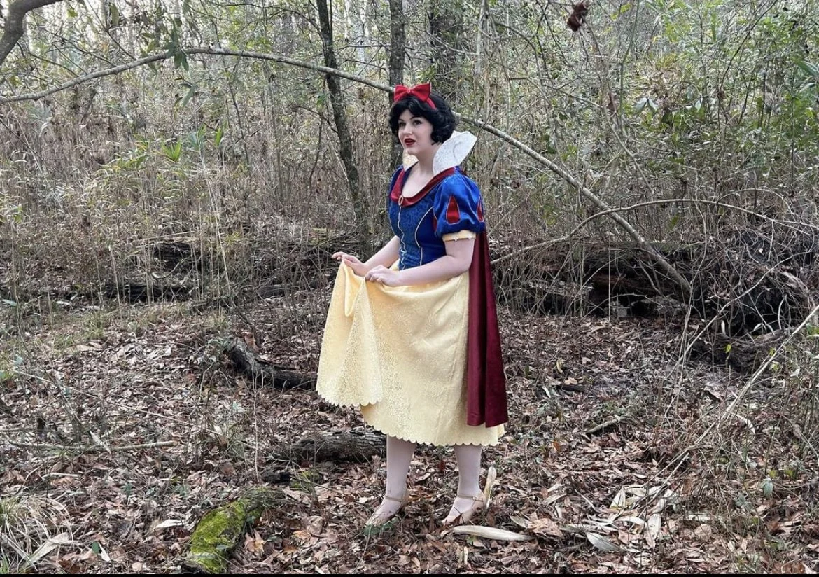 A woman dressed as Snow White stands in a wooded outdoors area with dry leaves and small plants on the ground, surrounded by trees and bushes.