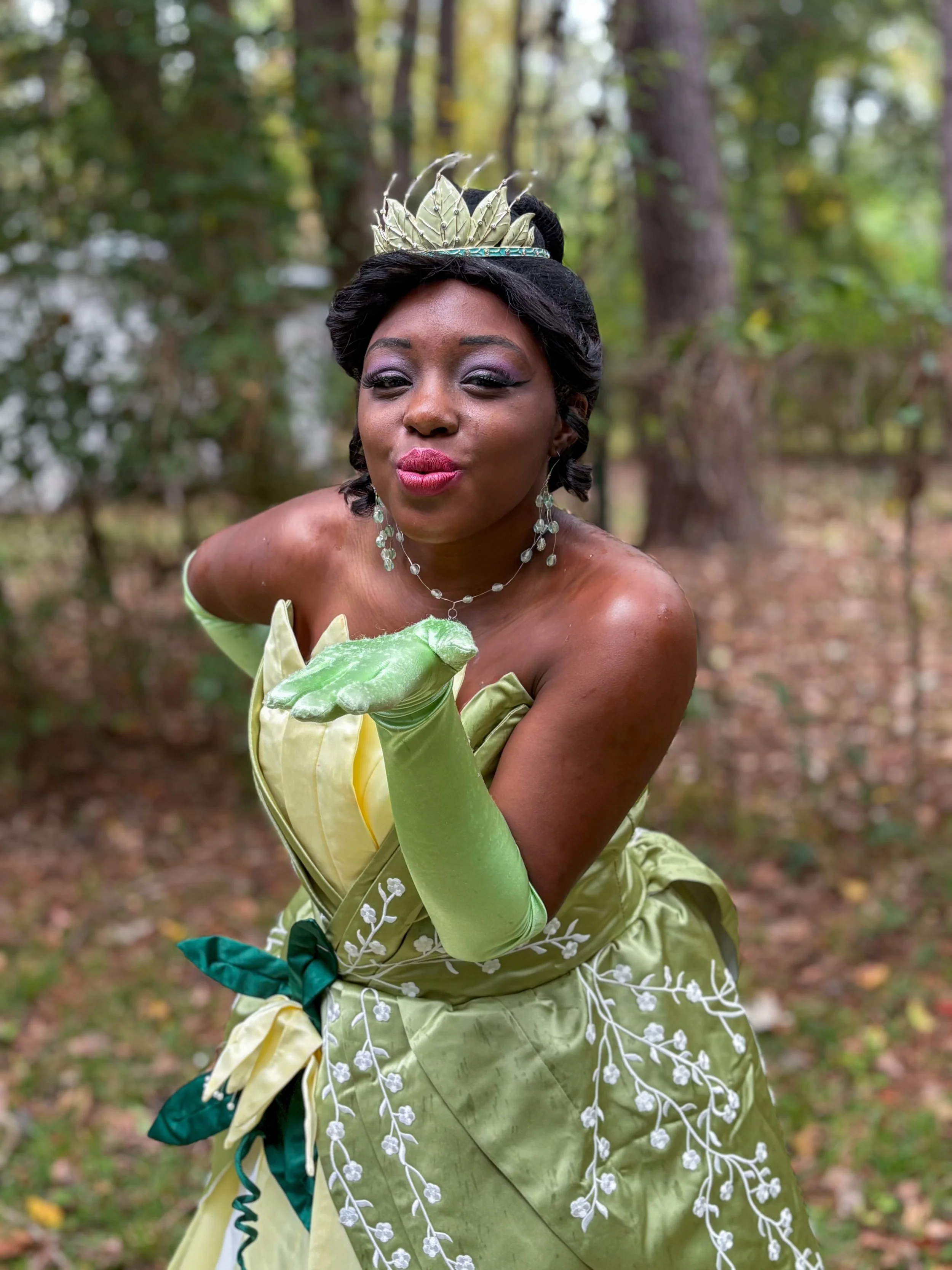 A woman is standing outdoors, dressed in a vintage-inspired dress with floral embroidery and long gloves, surrounded by lush greenery.