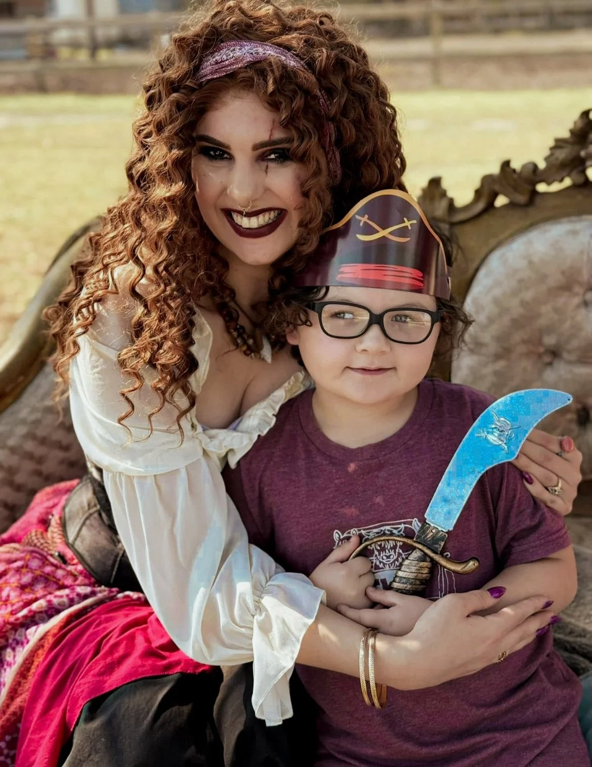 A woman with long curly red hair, wearing a purple headband, dark lipstick, and white blouse, is smiling and hugging a young boy who has curly hair, glasses, and is wearing a maroon T-shirt, a pirate hat, and holding a toy sword.