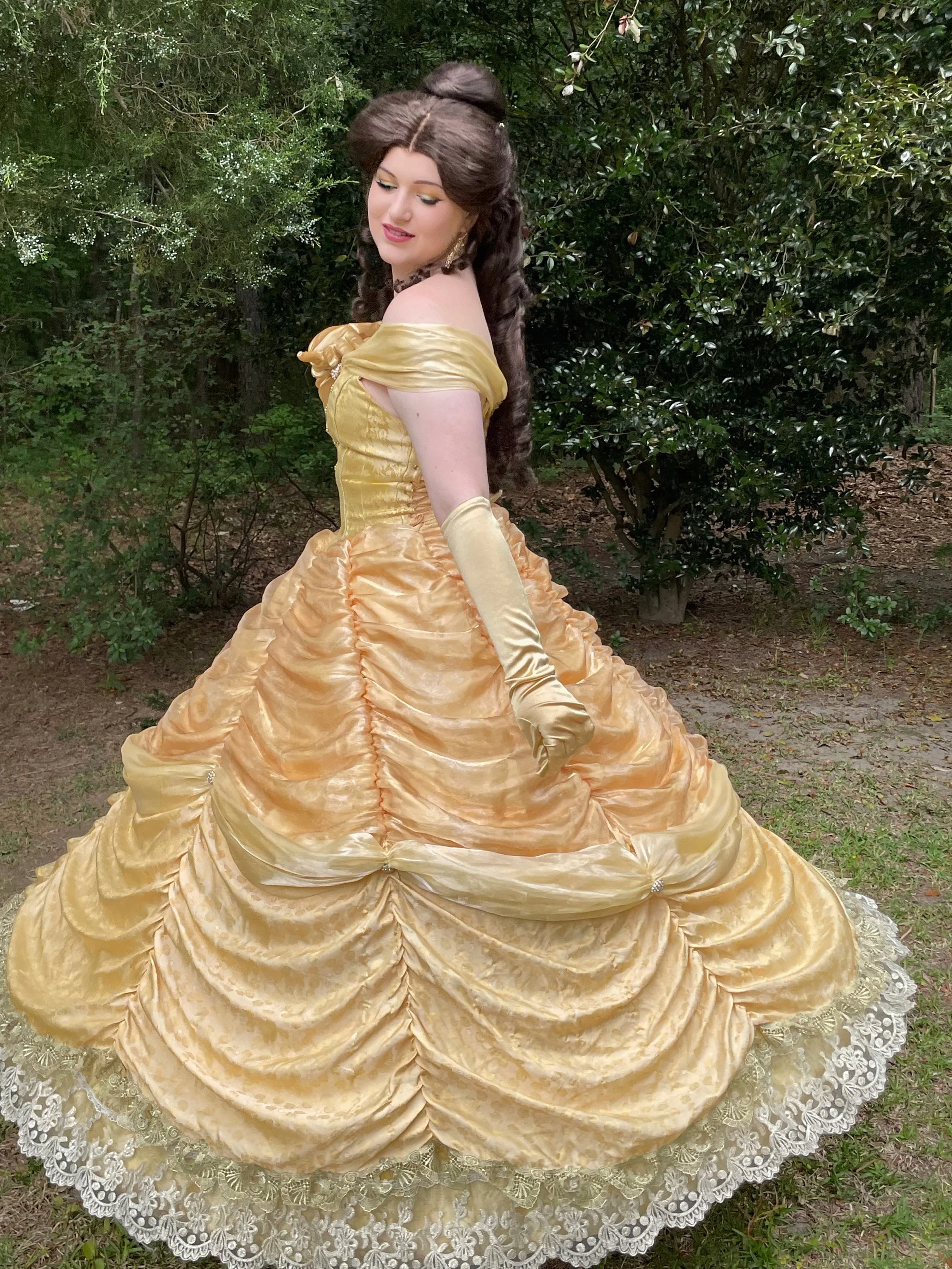 A woman in a Victorian-style yellow satin ball gown with puff sleeves, fitted bodice, and full skirt with ruched details and lace trim, standing outdoors surrounded by green foliage.