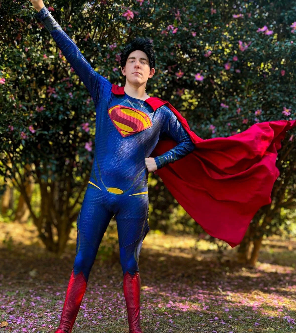 Person dressed as Superman standing outdoors, wearing a blue and red superhero costume with a cape, in front of a bush with pink flowers.