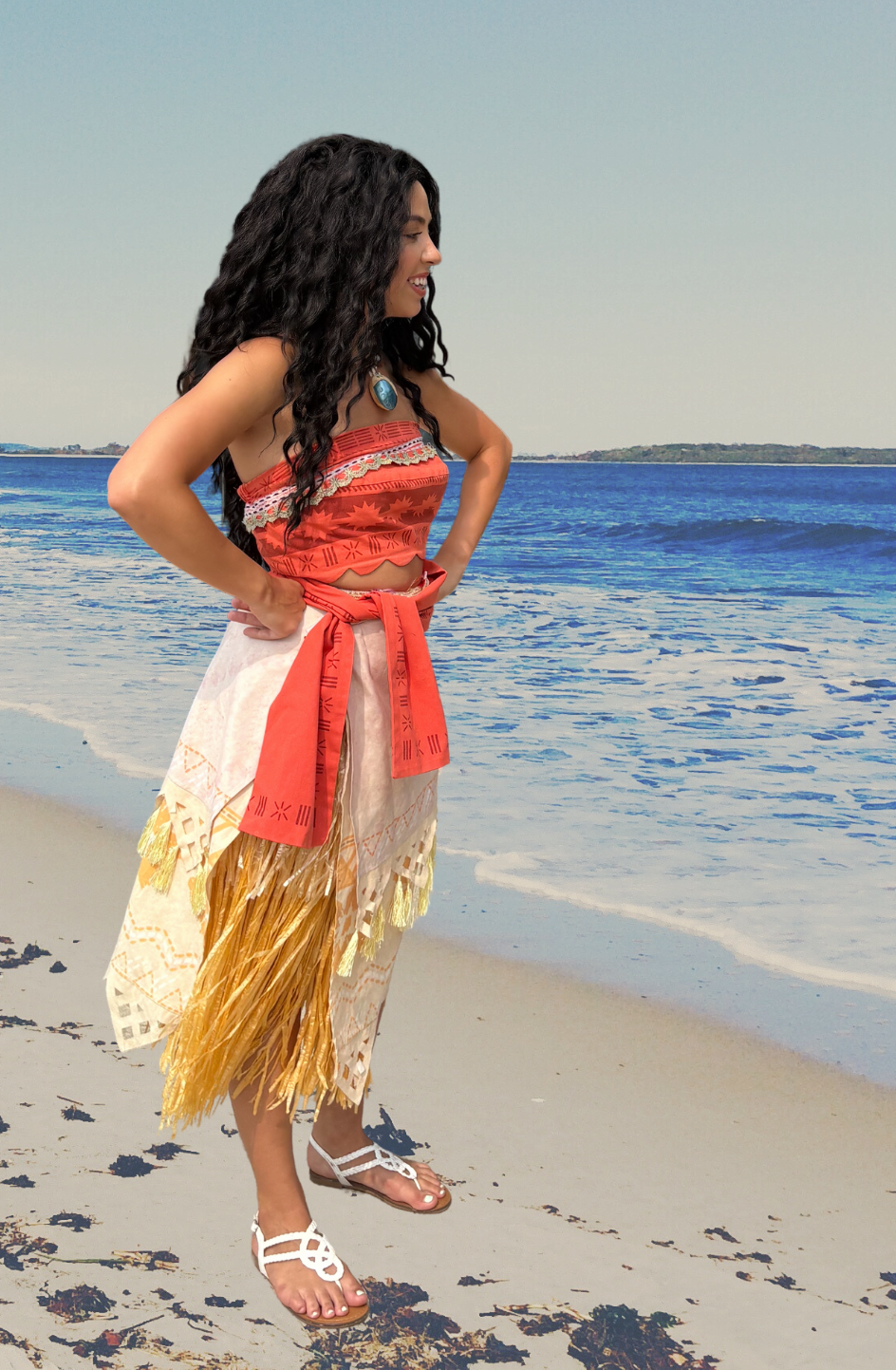A woman in traditional Hawaiian attire standing on a beach with the ocean in the background.