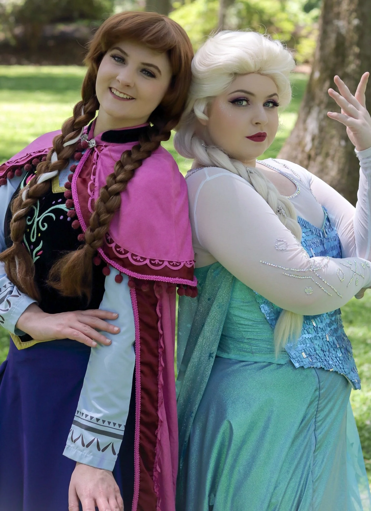 Two women in elaborate costumes, one with long red braided hair and the other with long platinum blonde braided hair, standing outdoors in front of trees, dressed as characters from Disney's Frozen.