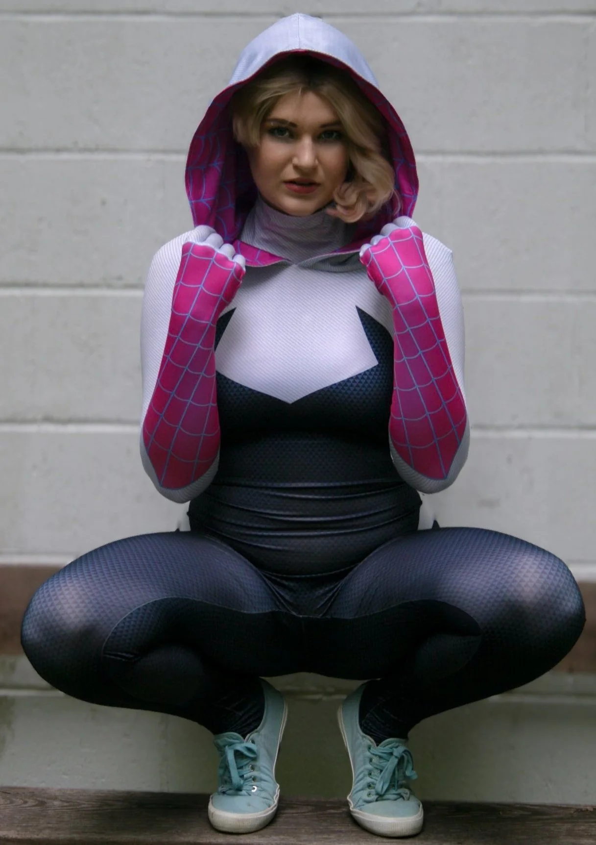 A woman dressed in a superhero costume, sitting in a squatting position against a concrete wall, wearing a Spiderman-themed outfit with a hood, gloves, leggings, and sneakers.