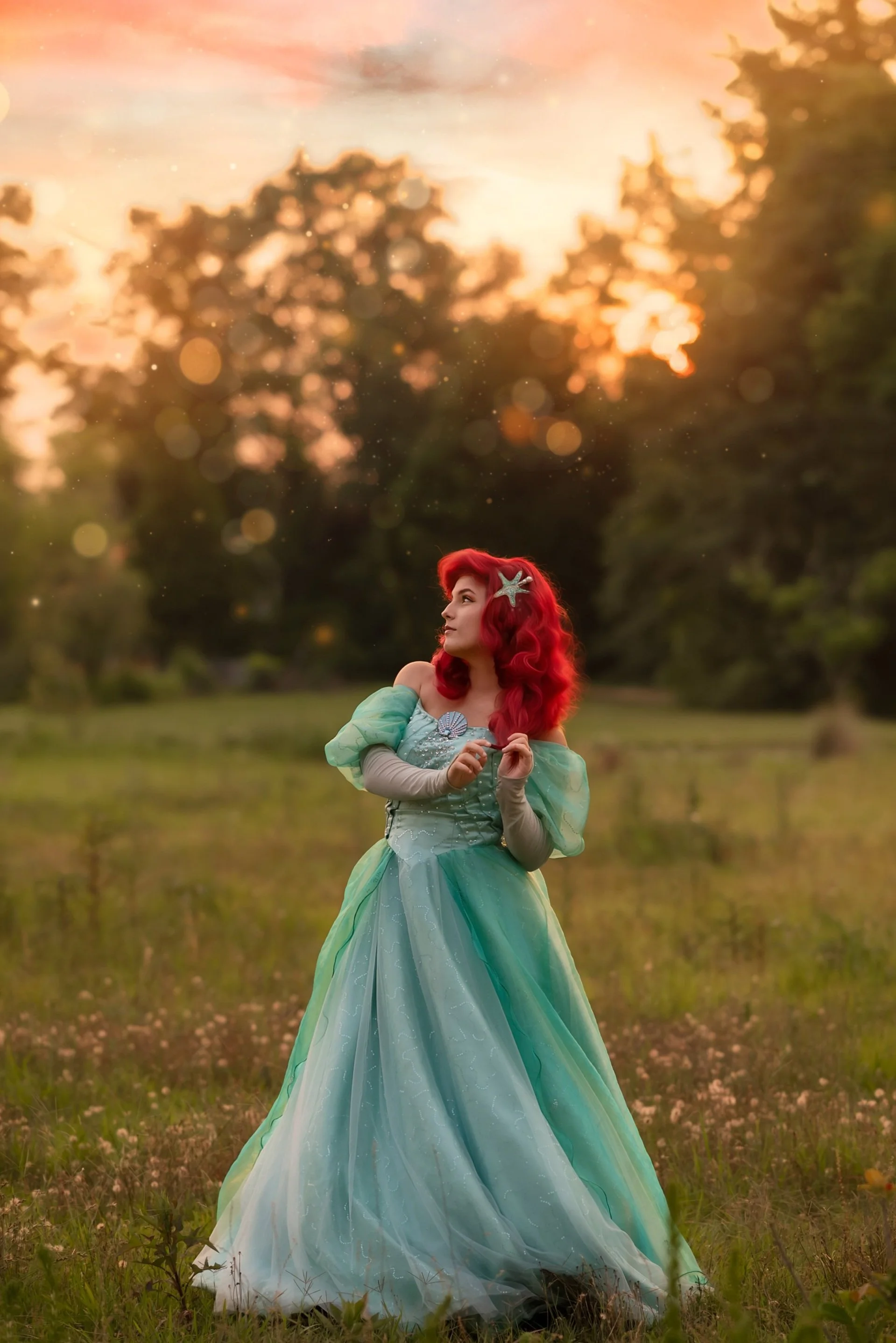 A woman with bright red hair dressed as a fairy or mermaid, wearing a flowing turquoise dress and a starfish hair accessory, standing in a grassy field at sunset with a bokeh effect in the background.
