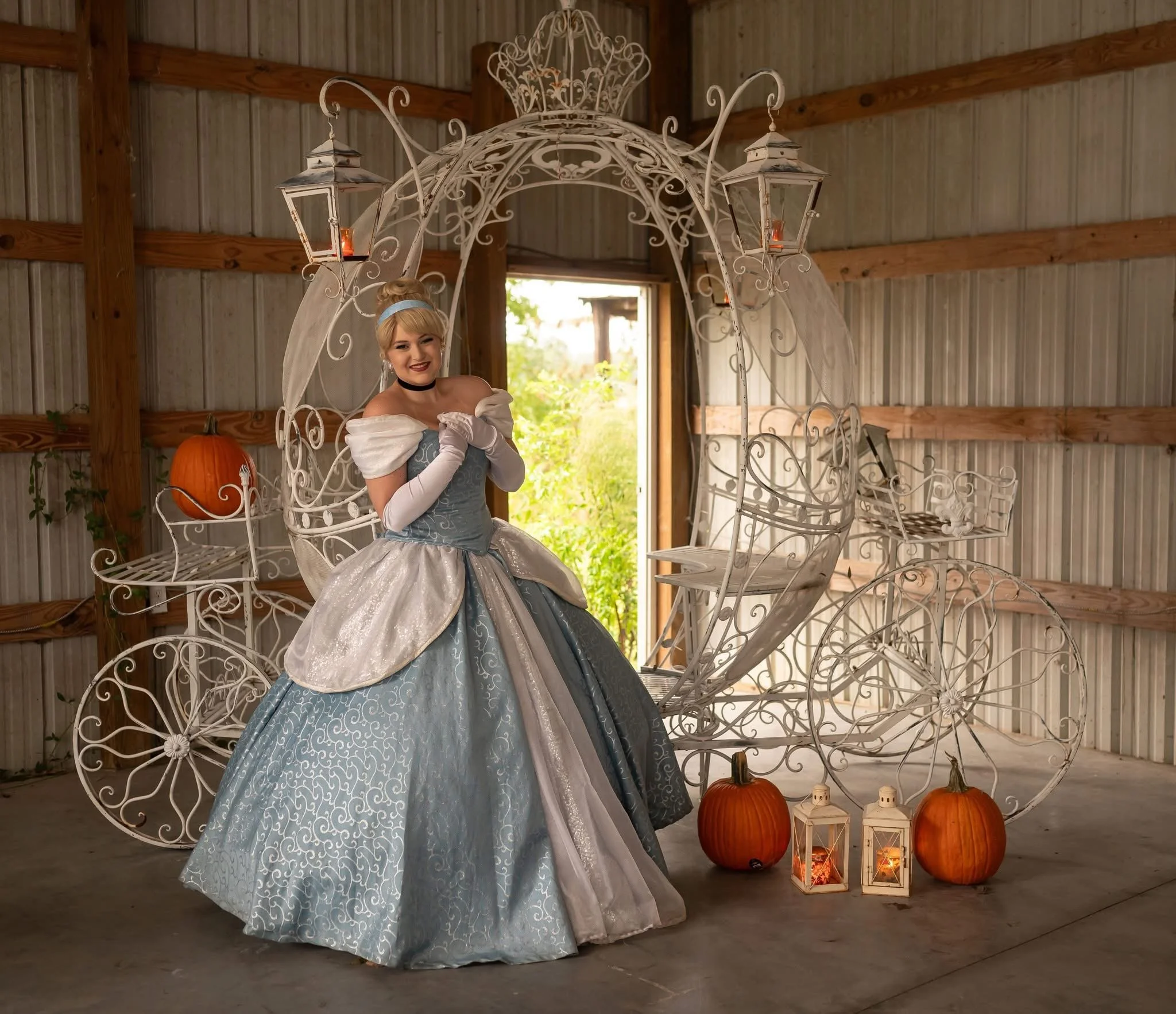 A woman dressed as Cinderella sitting on a decorative white metal carriage outdoors under a structure with lanterns, with greenery and trees in the background.