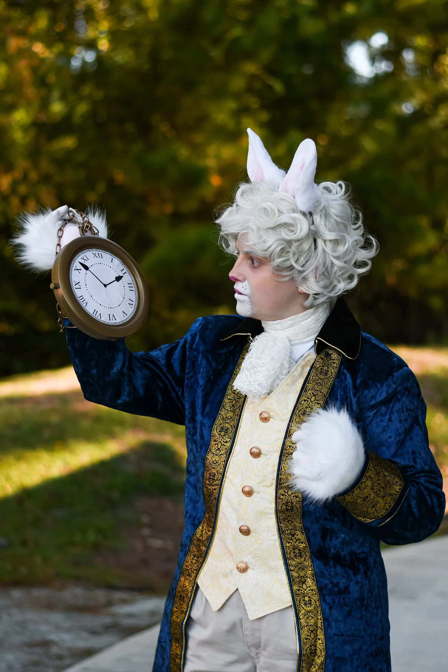 Person dressed as the White Rabbit from Alice in Wonderland, wearing a blue velvet jacket, cream vest, white ruffled cravat, and rabbit ears, holding a vintage clock outside with trees in the background.