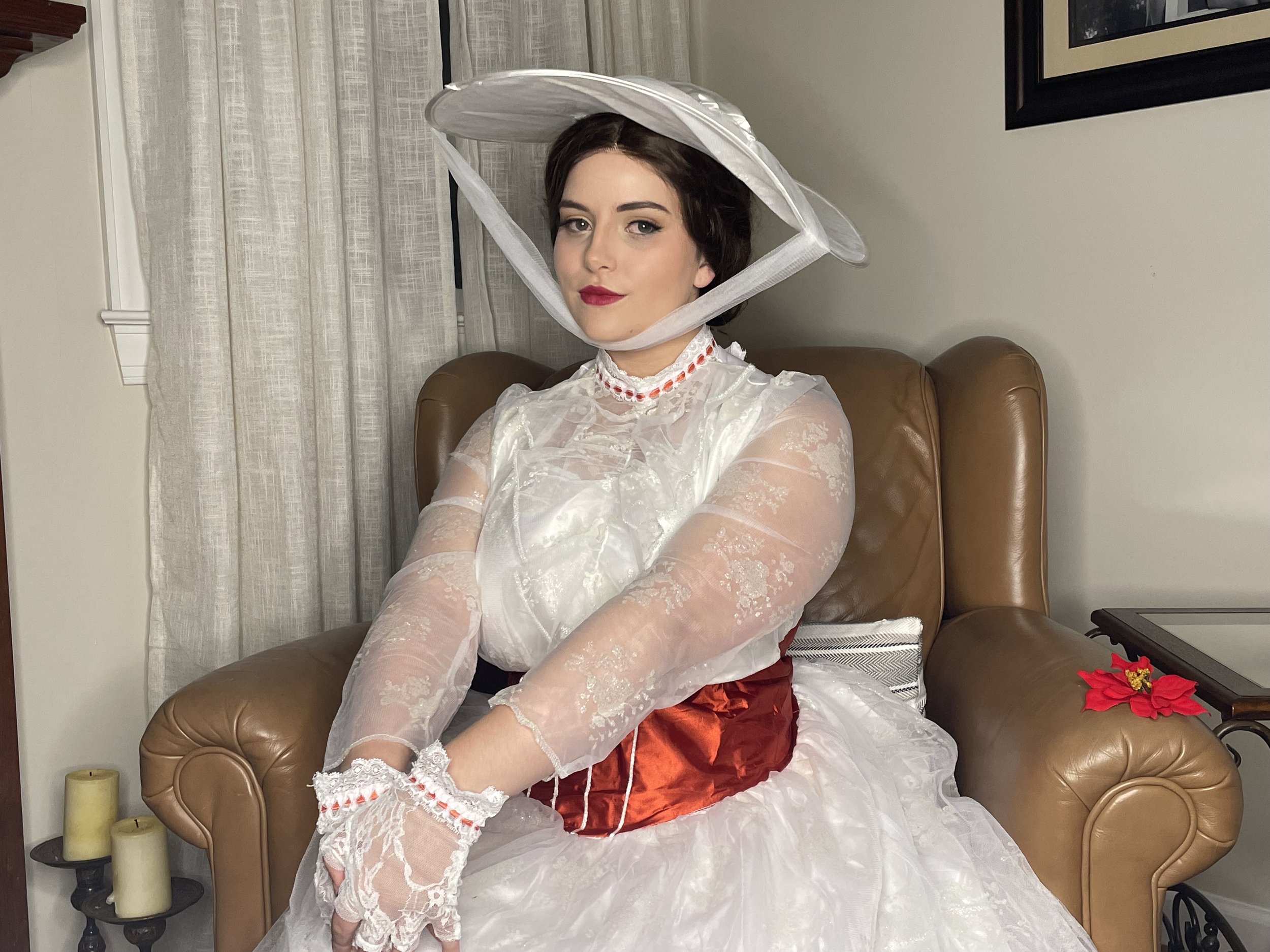 Woman dressed in vintage-style white lace dress with a sheer long-sleeve overlay, red corset, white petticoat, and a wide-brimmed white hat with netting, sitting on a brown leather armchair in a living room.