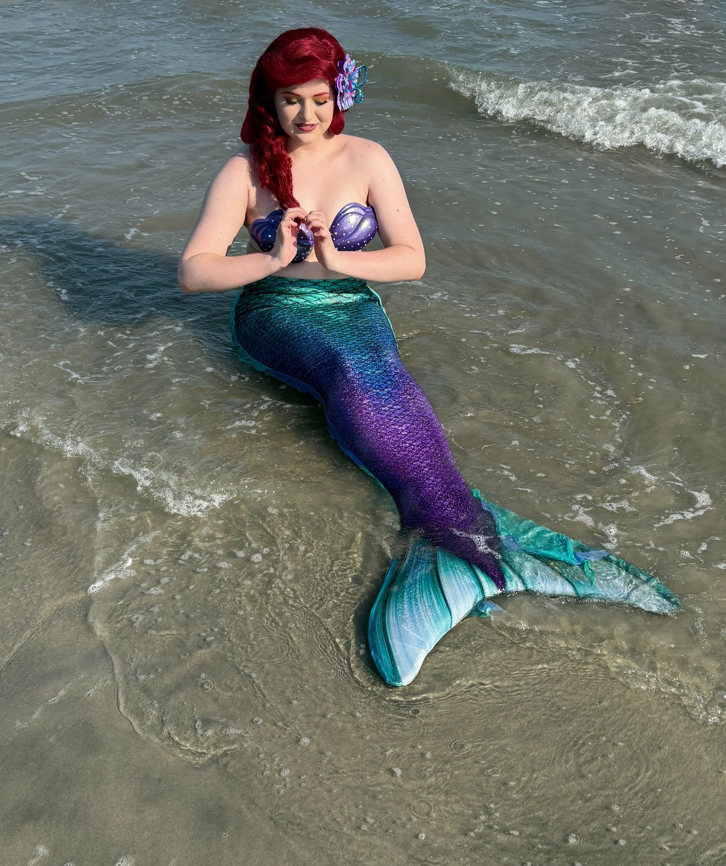 A woman with red hair dressed as a mermaid sitting in shallow ocean water.