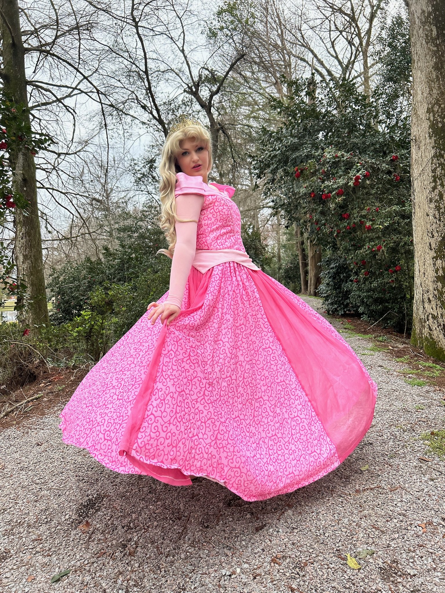 A woman dressed as a princess in a pink ball gown standing on a gravel path in a wooded area with trees and bushes, some with red berries.