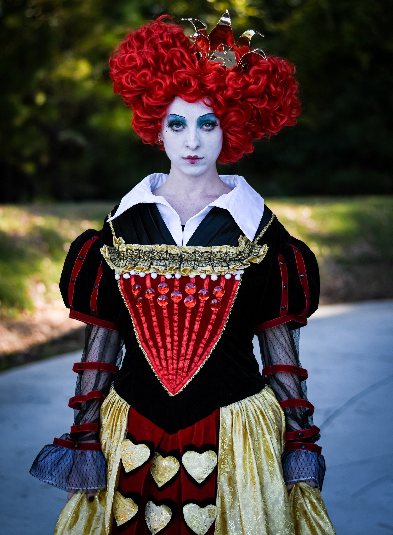 Person dressed as a queen of hearts with elaborate costume, red curly hair, white face makeup, and dramatic eye makeup, standing outdoors.