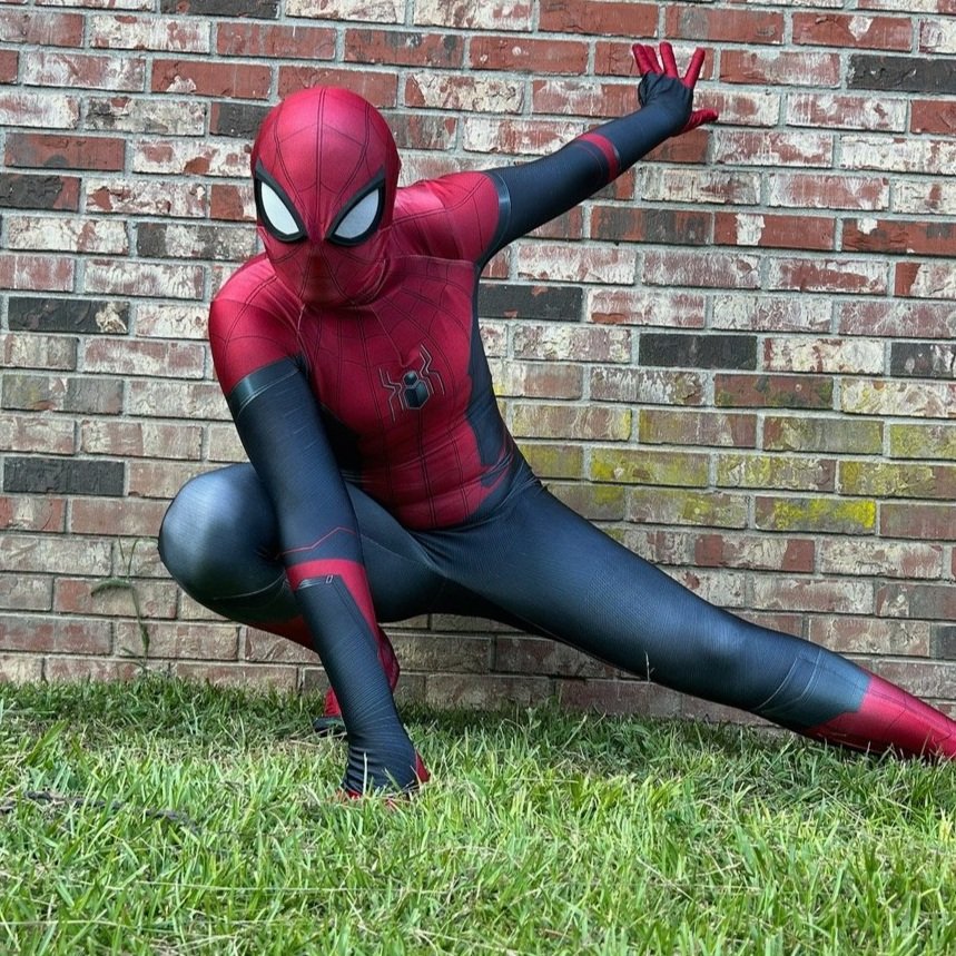 Person dressed as Spider-Man crouching on grass in front of brick wall, striking a dynamic pose.
