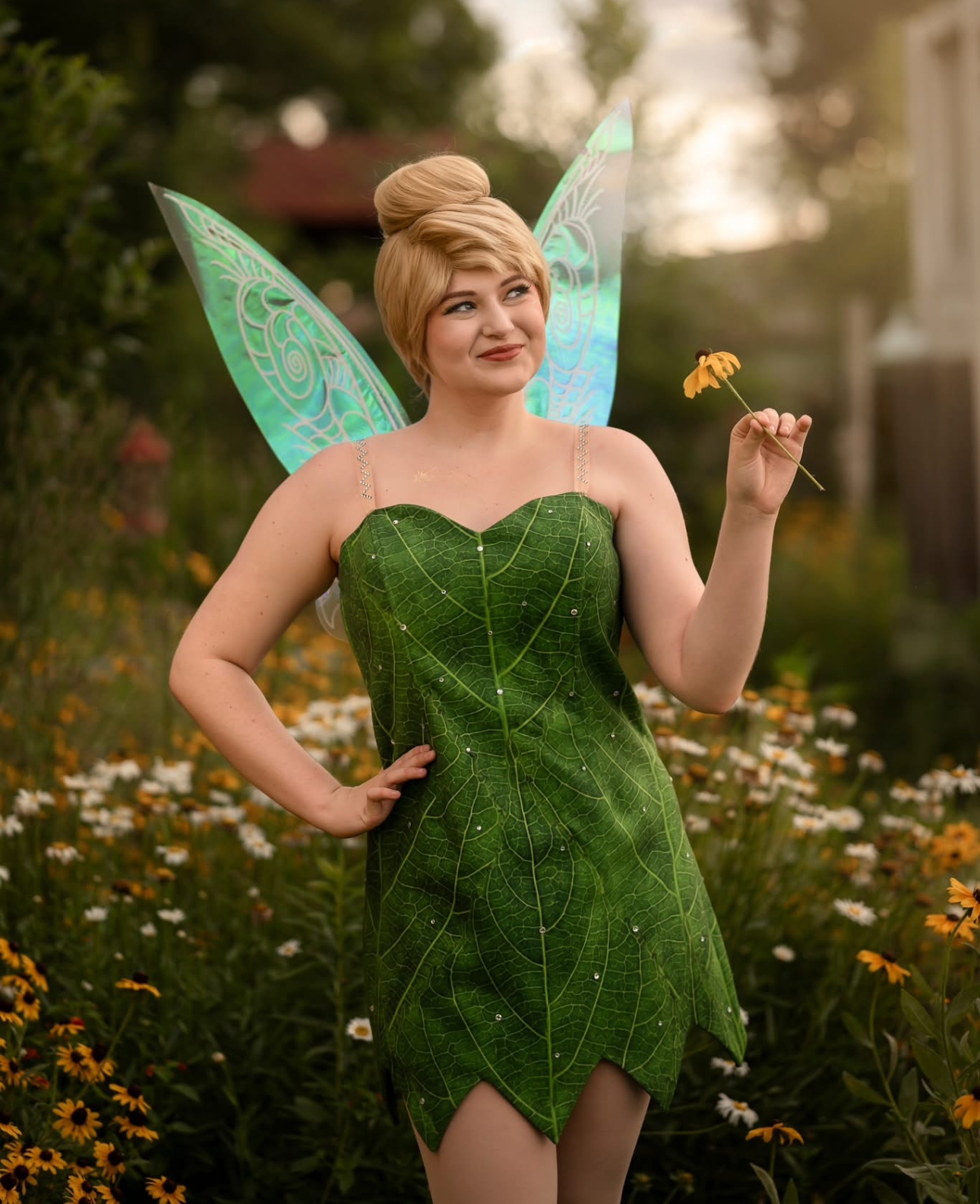 Person dressed as a fairy with leaf dress and iridescent fairy wings holding a yellow flower in a garden of daisies.