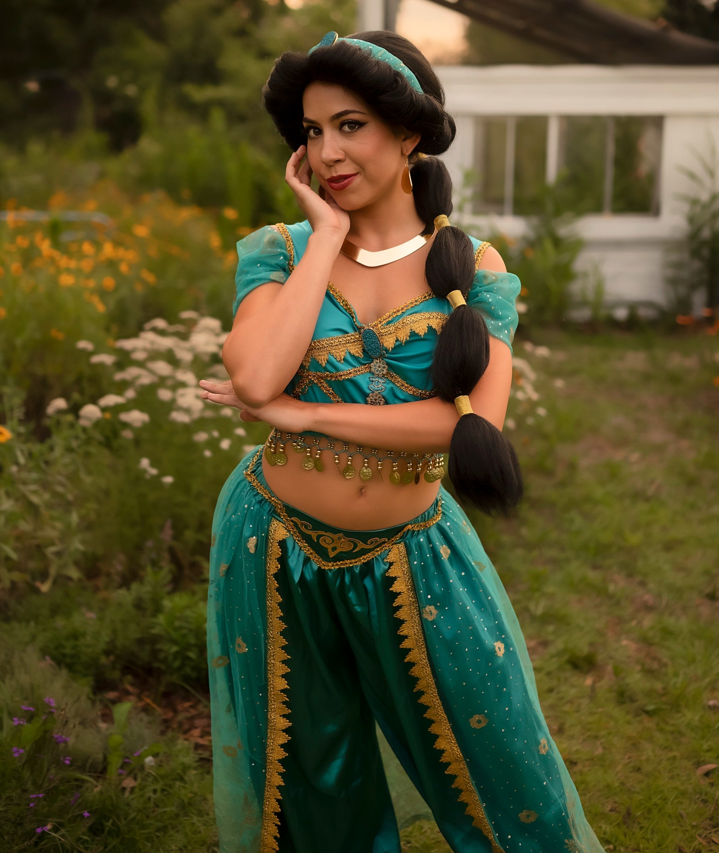 Woman in traditional turquoise costume with gold accents, standing outdoors in a garden at sunset.