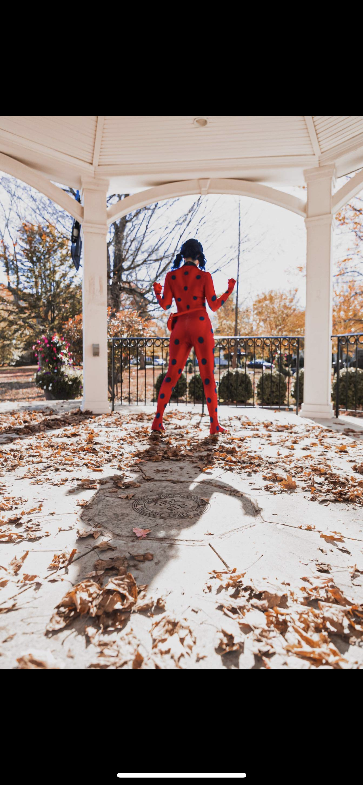 Person wearing a red and black polka-dot jumpsuit standing on a covered porch, facing away, with autumn leaves on the ground and trees in the background.