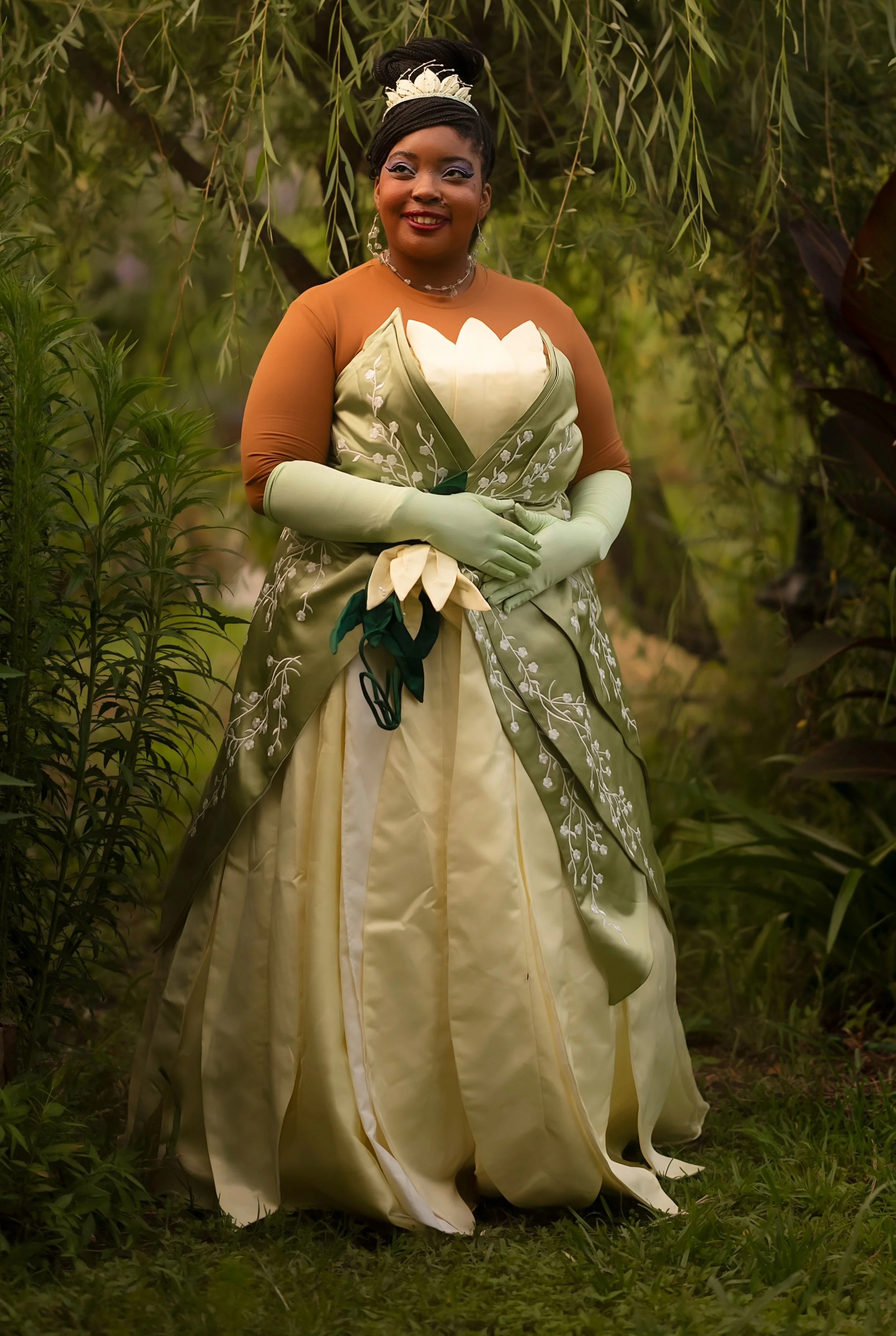 A woman is standing outdoors, dressed in a vintage-inspired dress with floral embroidery and long gloves, surrounded by lush greenery.