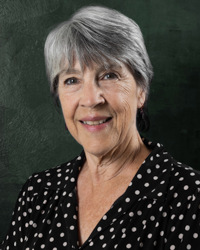 A smiling older woman with short gray hair, wearing a black blouse with white polka dots, standing in front of a dark green background.