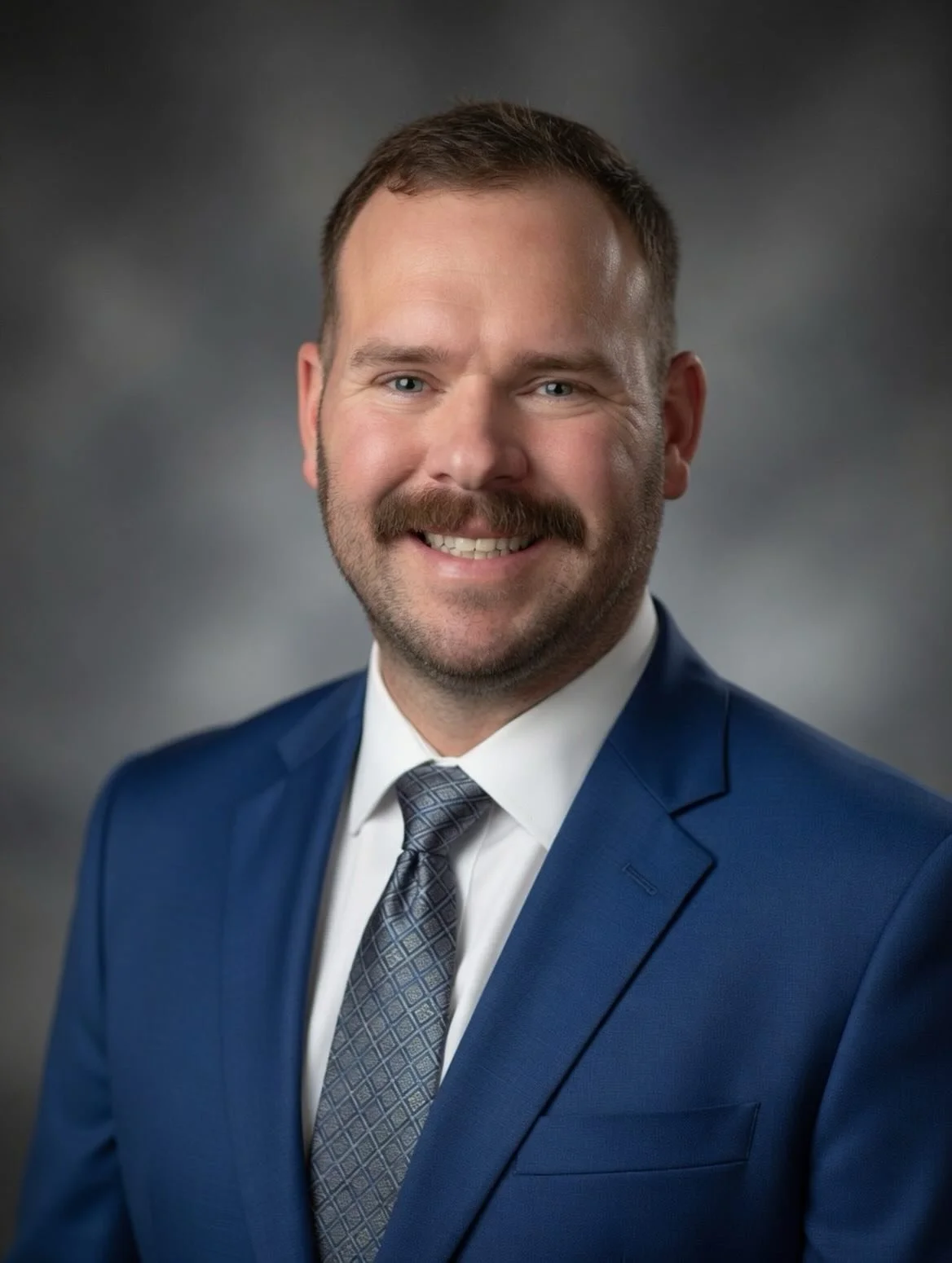A professional headshot of a man with short brown hair, a beard, and a mustache, wearing a blue suit, white shirt, and patterned tie, smiling against a dark gray background.