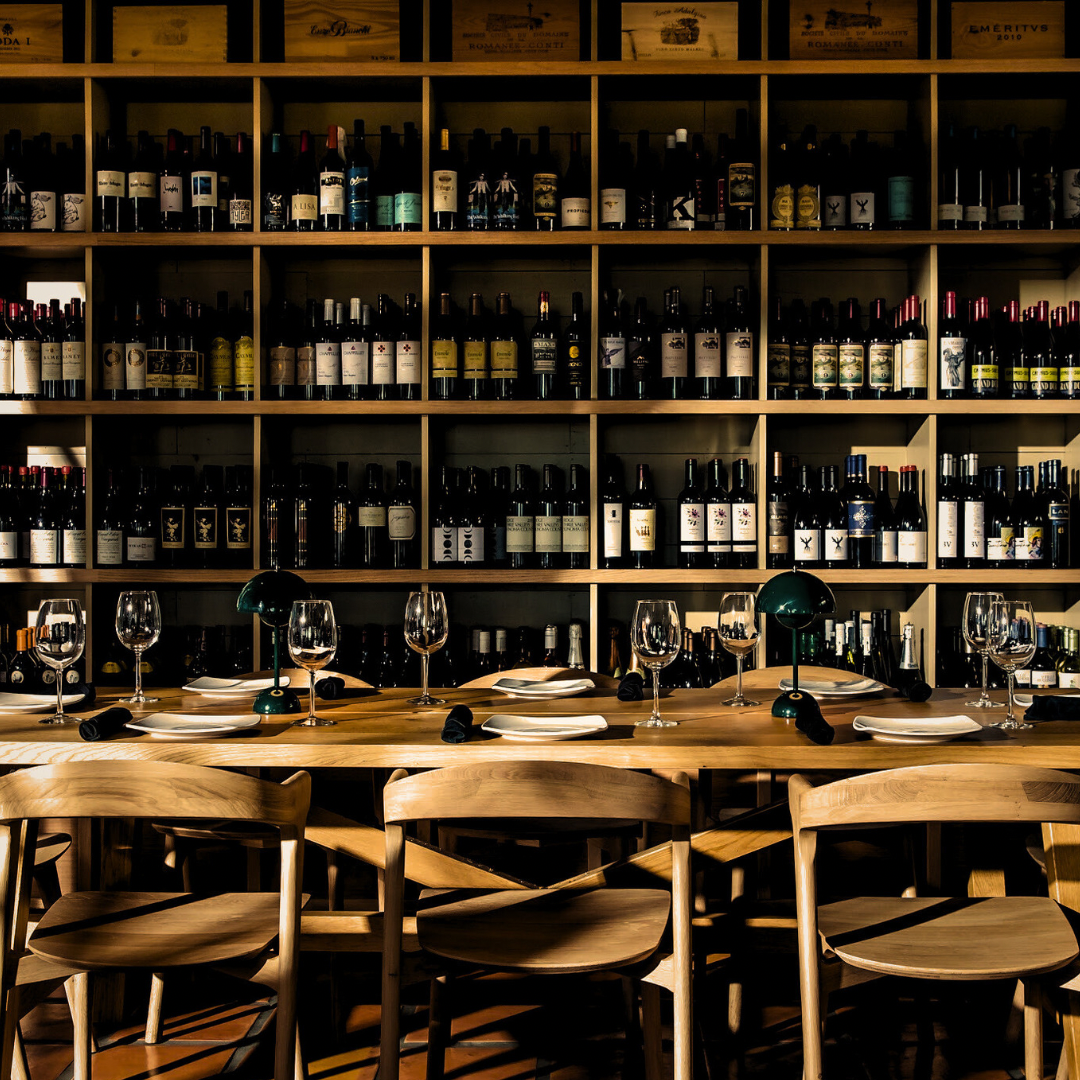 Long communal table set with wine glasses in front of a floor-to-ceiling wall of wine bottles
