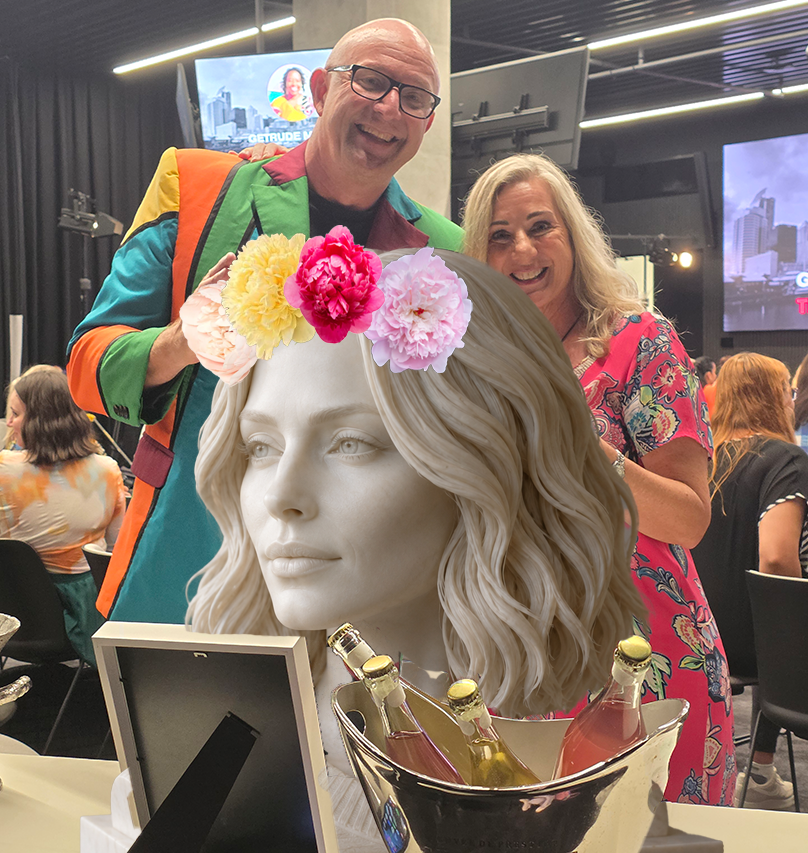 A man and woman smiling at a gathering, with a sculpture of a woman's head with flowers on her hair and bottles in a bucket in front of them.