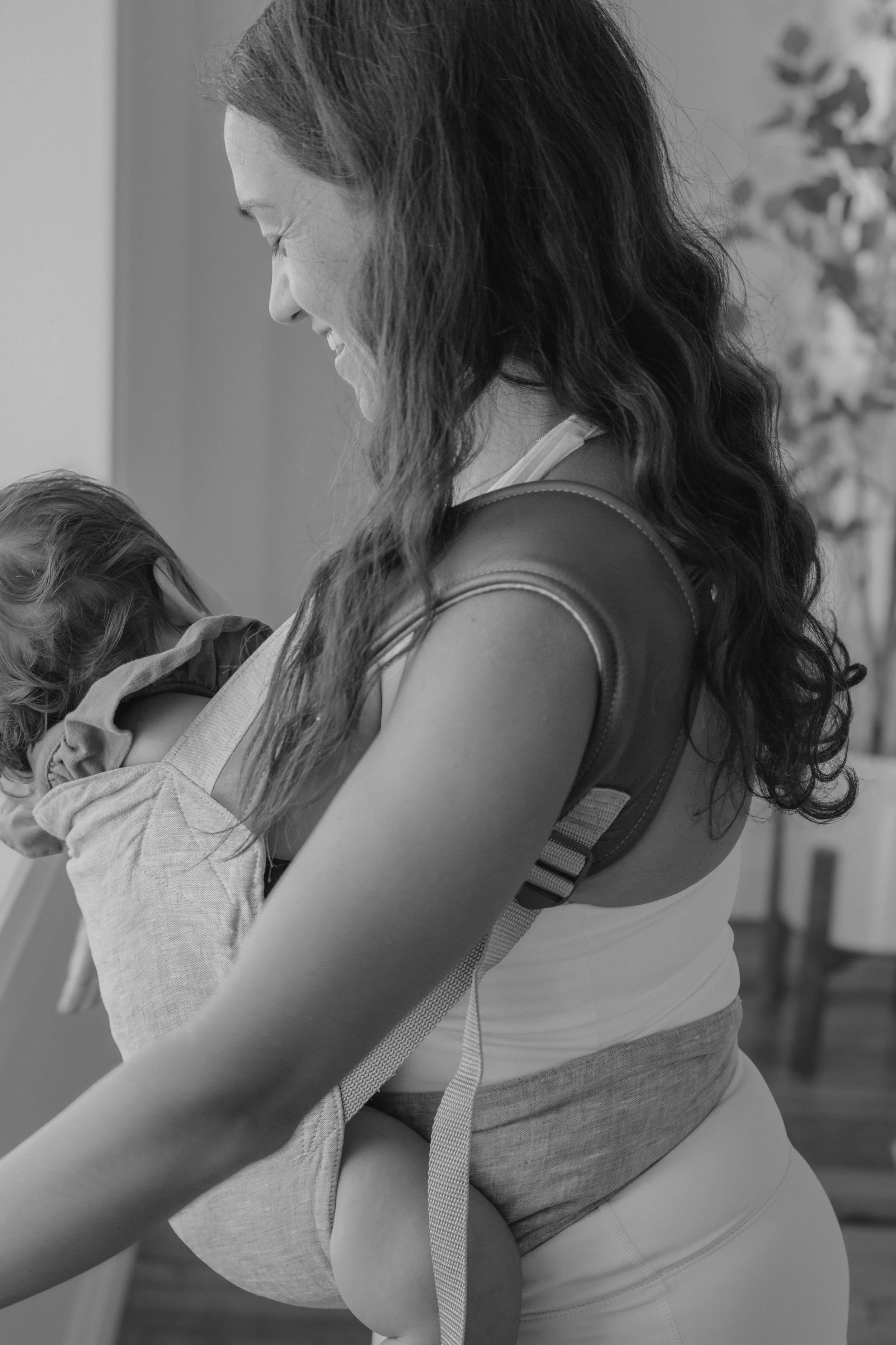 A woman with long wavy hair smiling while holding a child in her arms, both looking downward, in black and white.