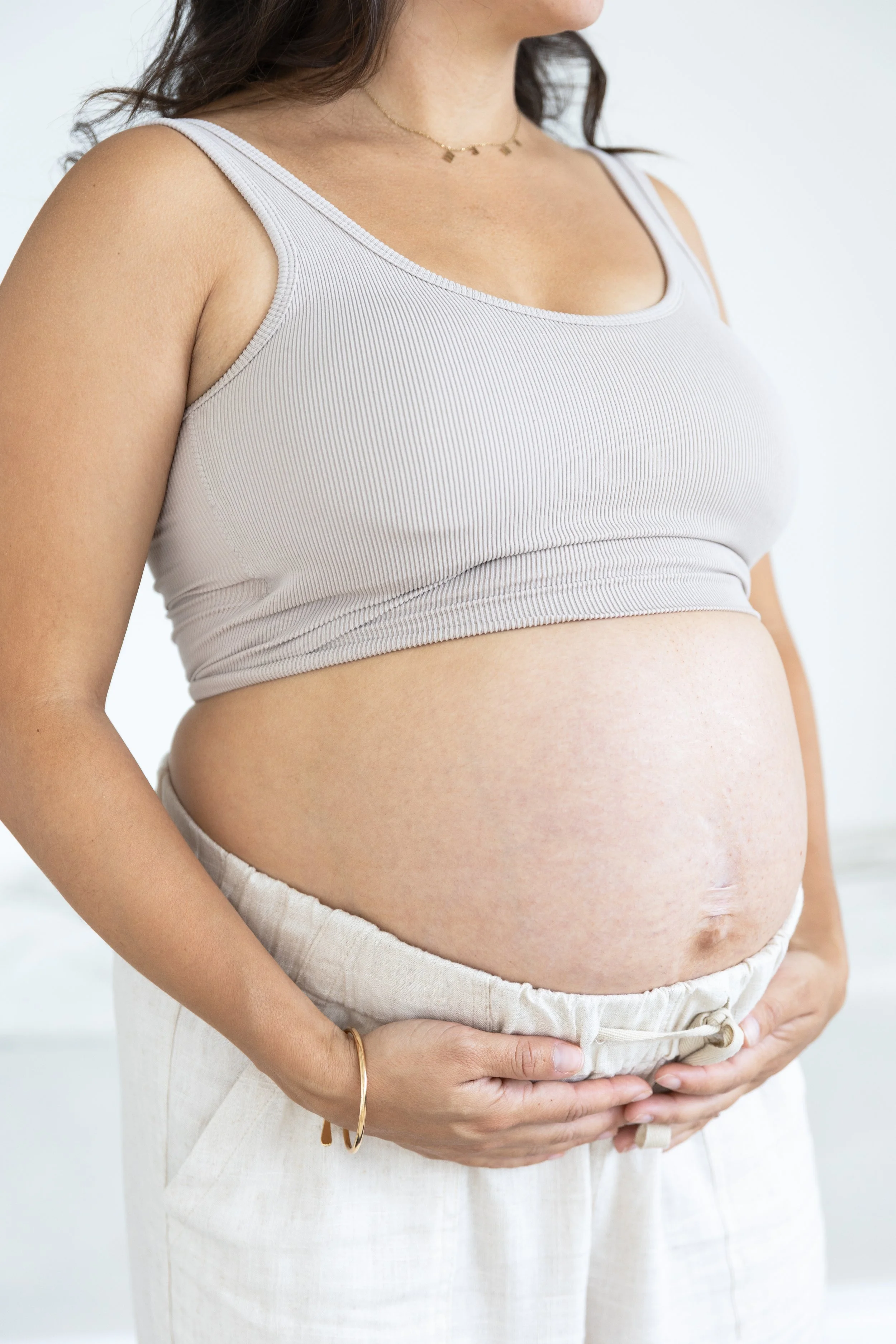 A pregnant woman wearing a beige sports bra and light-colored loose pants, holding her belly with both hands.