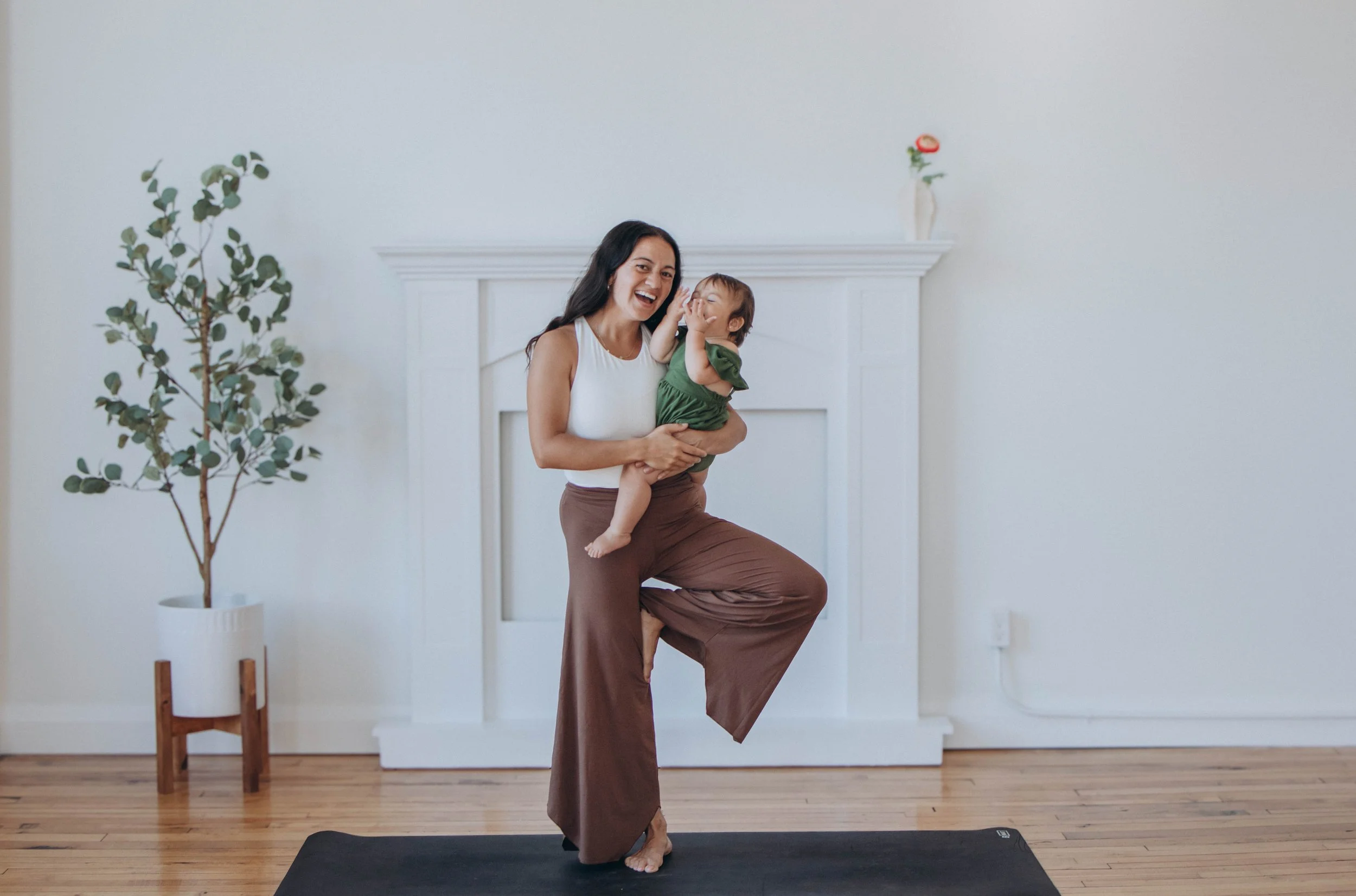 A woman holding a laughing child in a living room, with a white fireplace, green potted plant, and minimal decor.