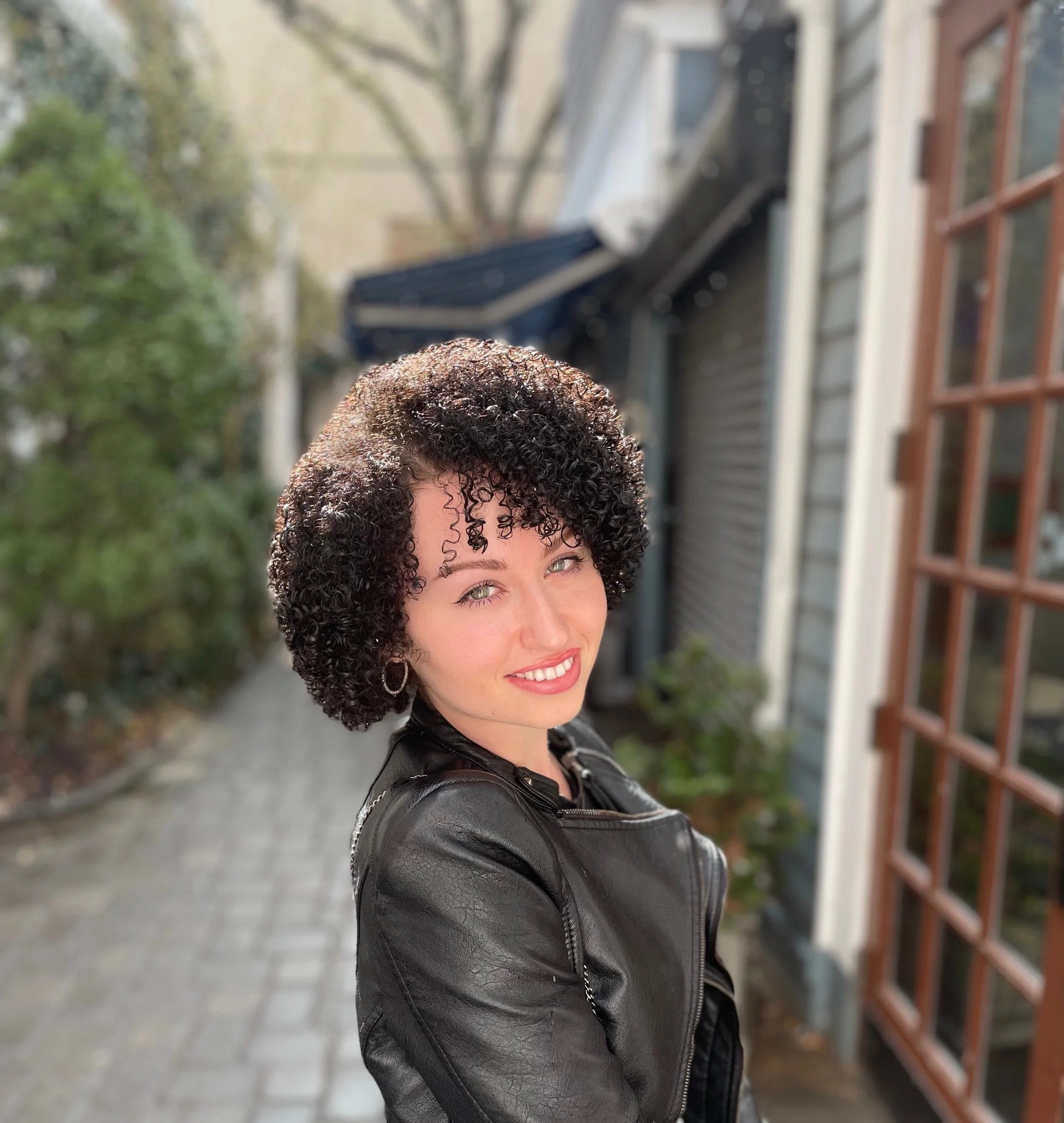 A woman with curly black hair and blue eyes smiling outdoors in front of a building with a wooden door and windows, wearing a black leather jacket.
