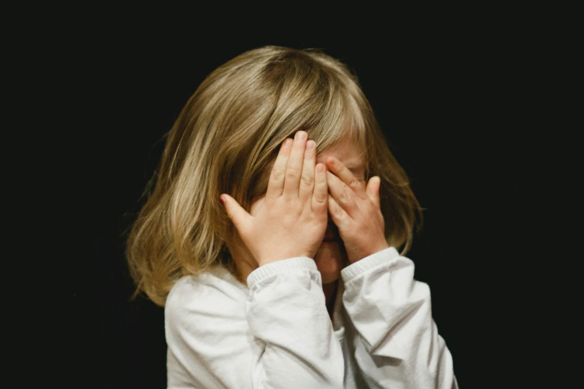 A young girl with blonde hair covering her face with her hands, appearing to be upset or crying, against a black background.