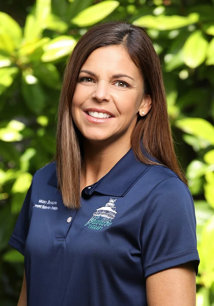 A woman with brown hair and a big smile, wearing a navy blue polo shirt with a logo, standing outdoors with green leaves in the background.
