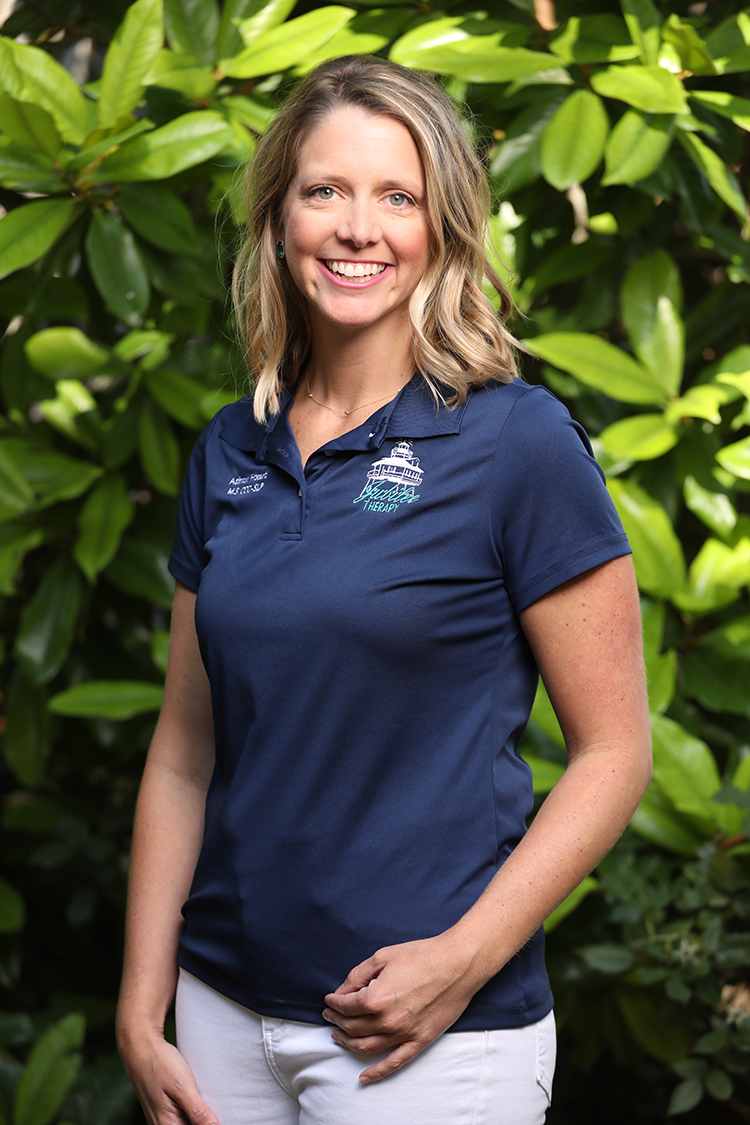 A smiling woman with blonde hair, wearing a navy blue polo shirt with a logo, standing outdoors in front of green foliage.