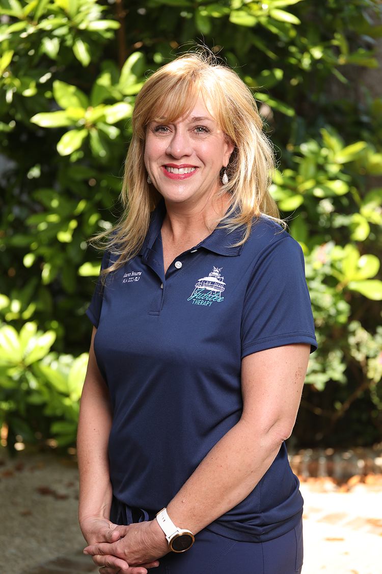 A woman with blonde hair wearing a navy blue polo shirt with a logo that says 'Aquatic Therapy' on it, standing outdoors with green foliage in the background.