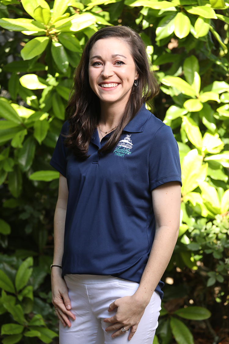 Smiling woman with long brown hair, wearing a blue polo shirt and white pants, standing outdoors in front of green leafy plants.