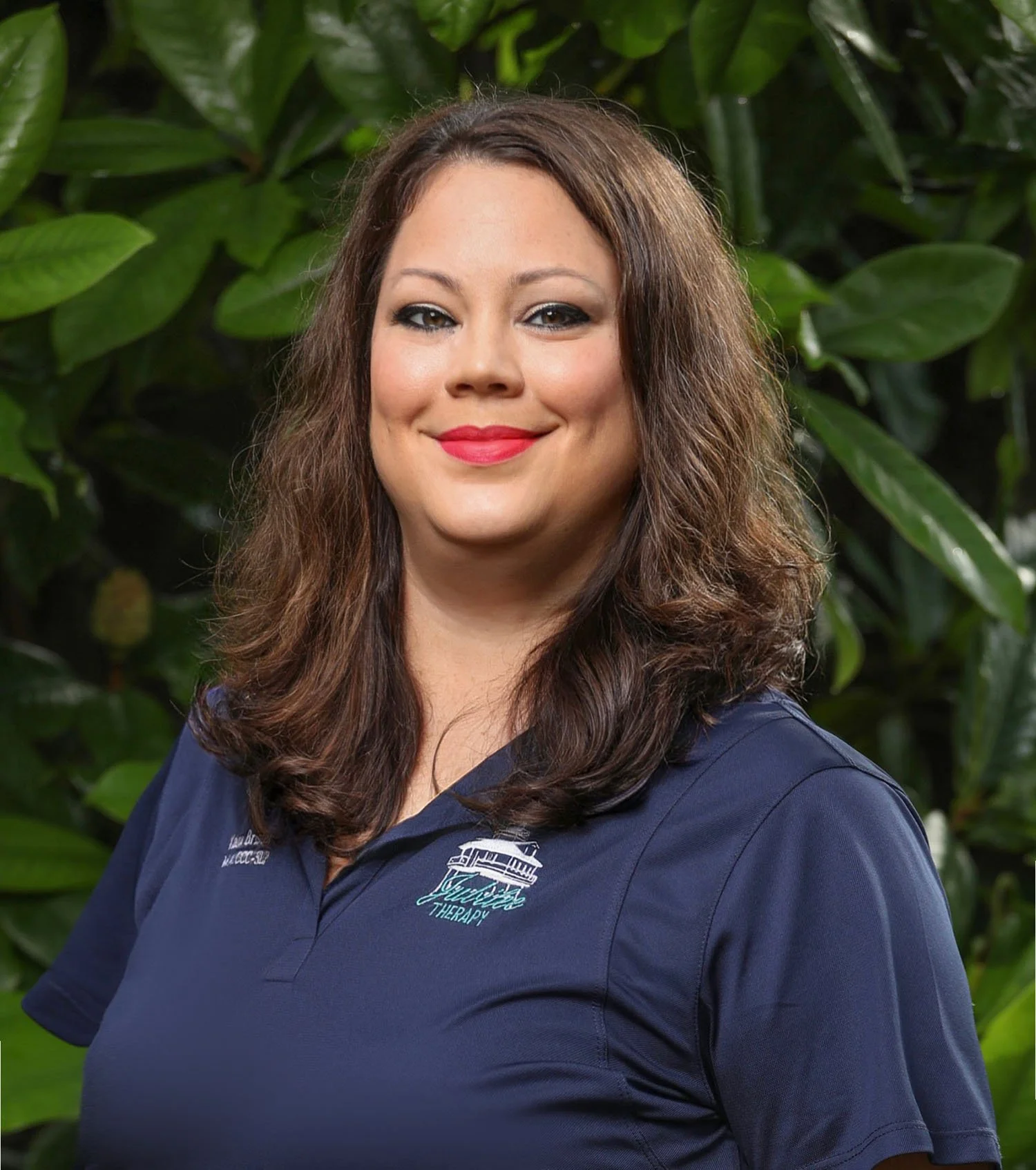 A woman with long brown hair, wearing a navy blue polo shirt with a logo, standing outdoors in front of leafy green plants.