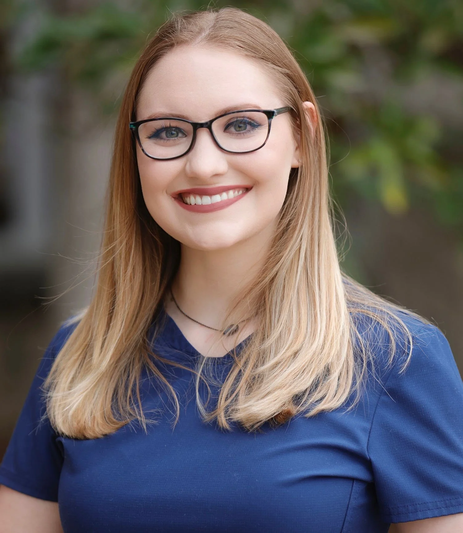 A young woman with long blonde hair, wearing glasses and a blue shirt, smiling outdoors with blurred green foliage in the background.