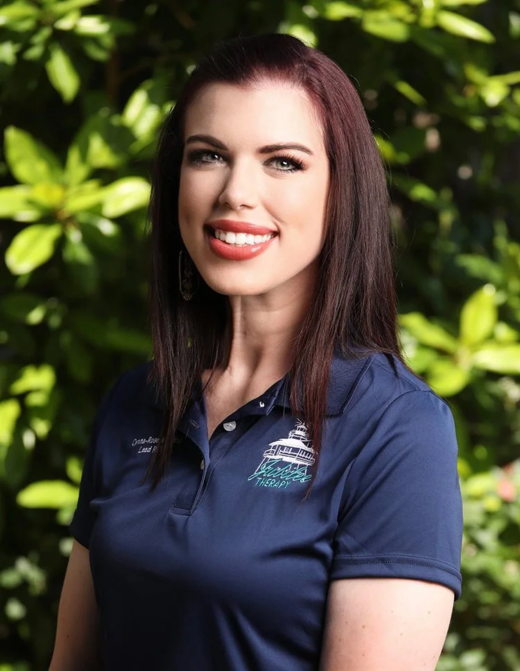 A woman with long dark hair smiling outdoors, wearing a navy polo shirt with a logo and text on it, surrounded by green foliage.
