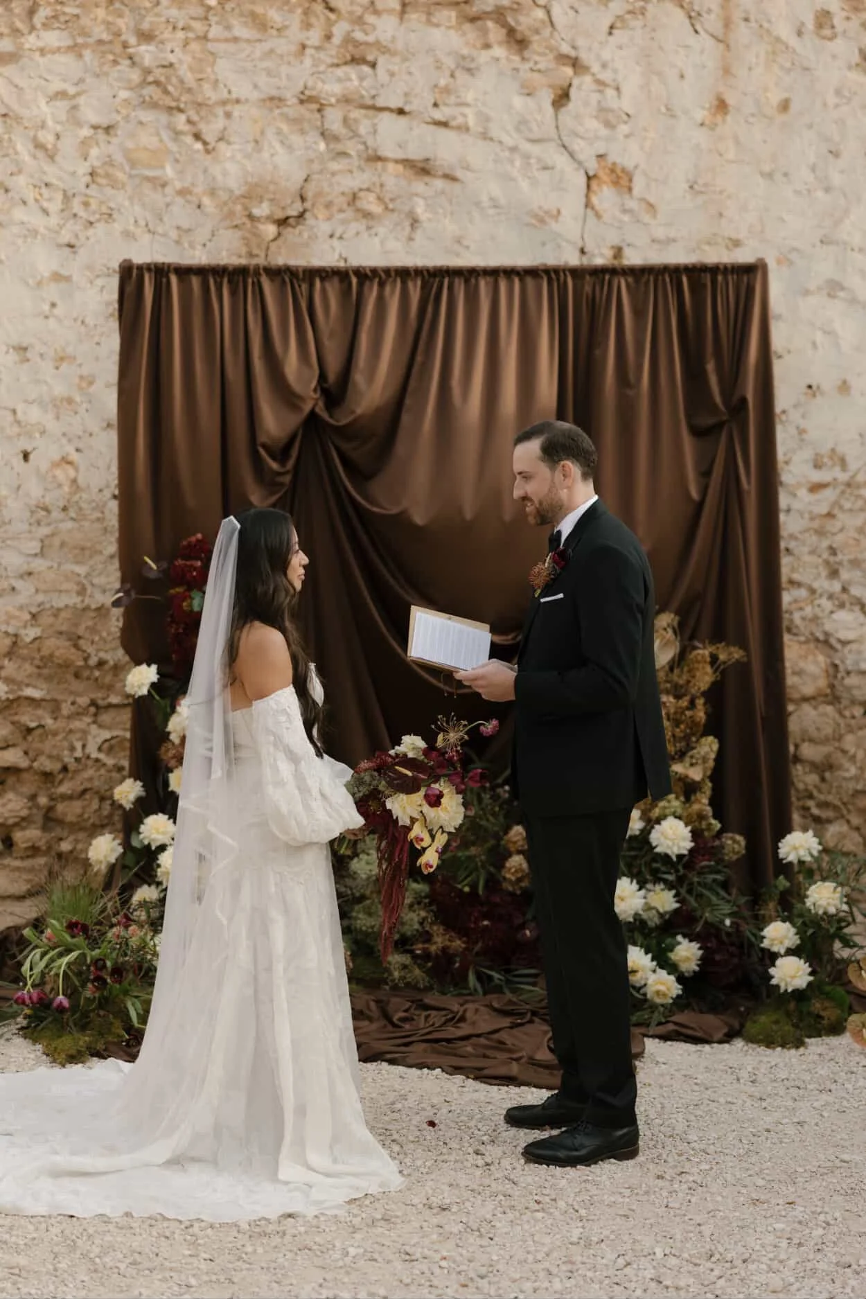 A happy couple outdoors, with the woman giving a piggyback ride on the man's back, holding a pink flower, surrounded by green trees.