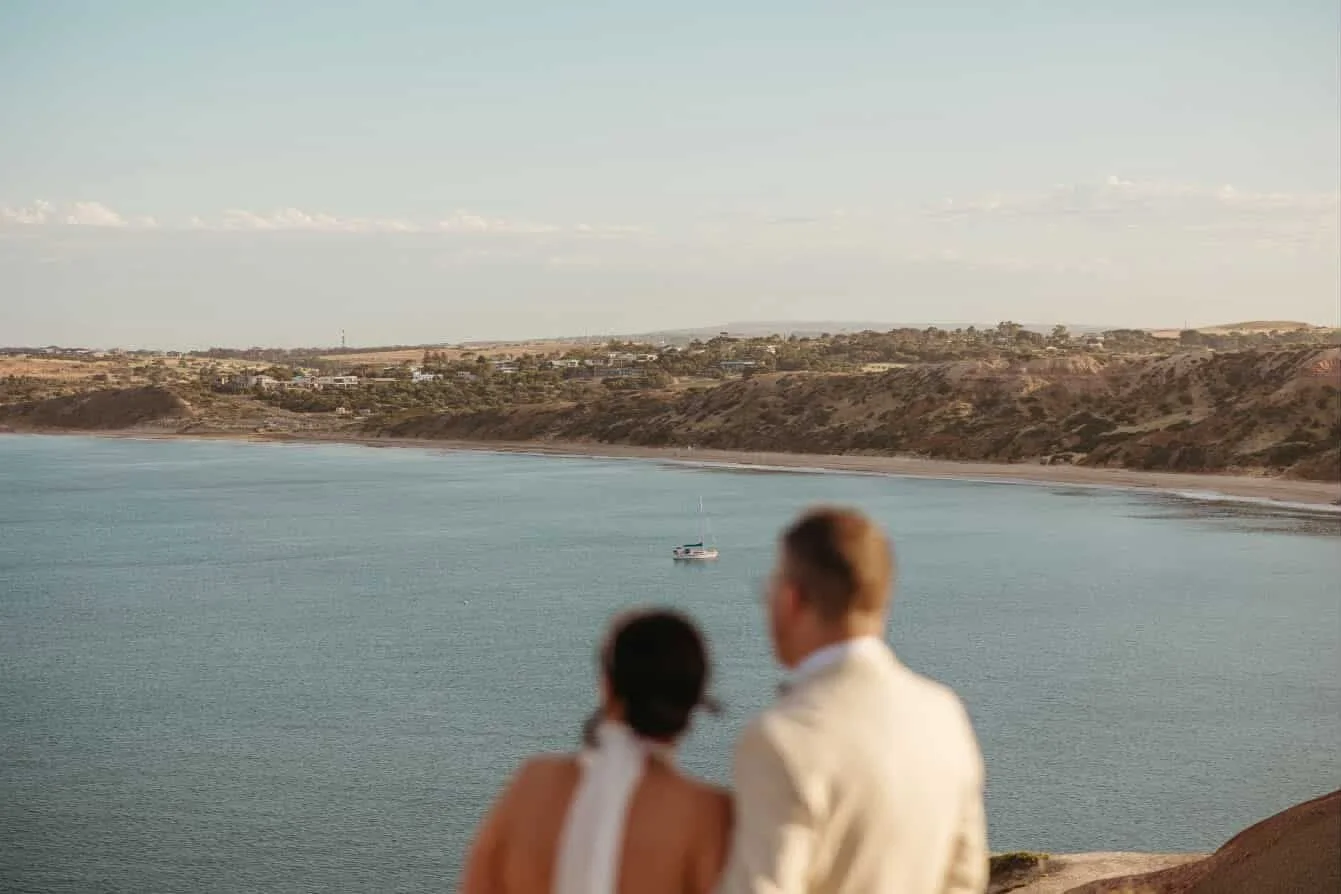 A happy couple outdoors, with the woman giving a piggyback ride on the man's back, holding a pink flower, surrounded by green trees.