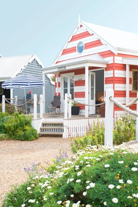 Colorful beach house with red and white stripes, small porch, and garden with white daisies.