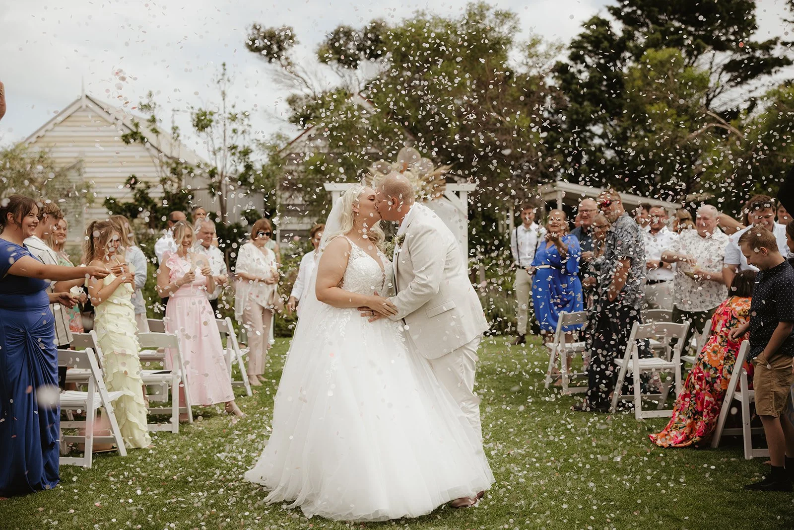 A newlywed couple holding hands and smiling as they walk through a shower of confetti during their wedding ceremony in a lush, indoor garden setting.