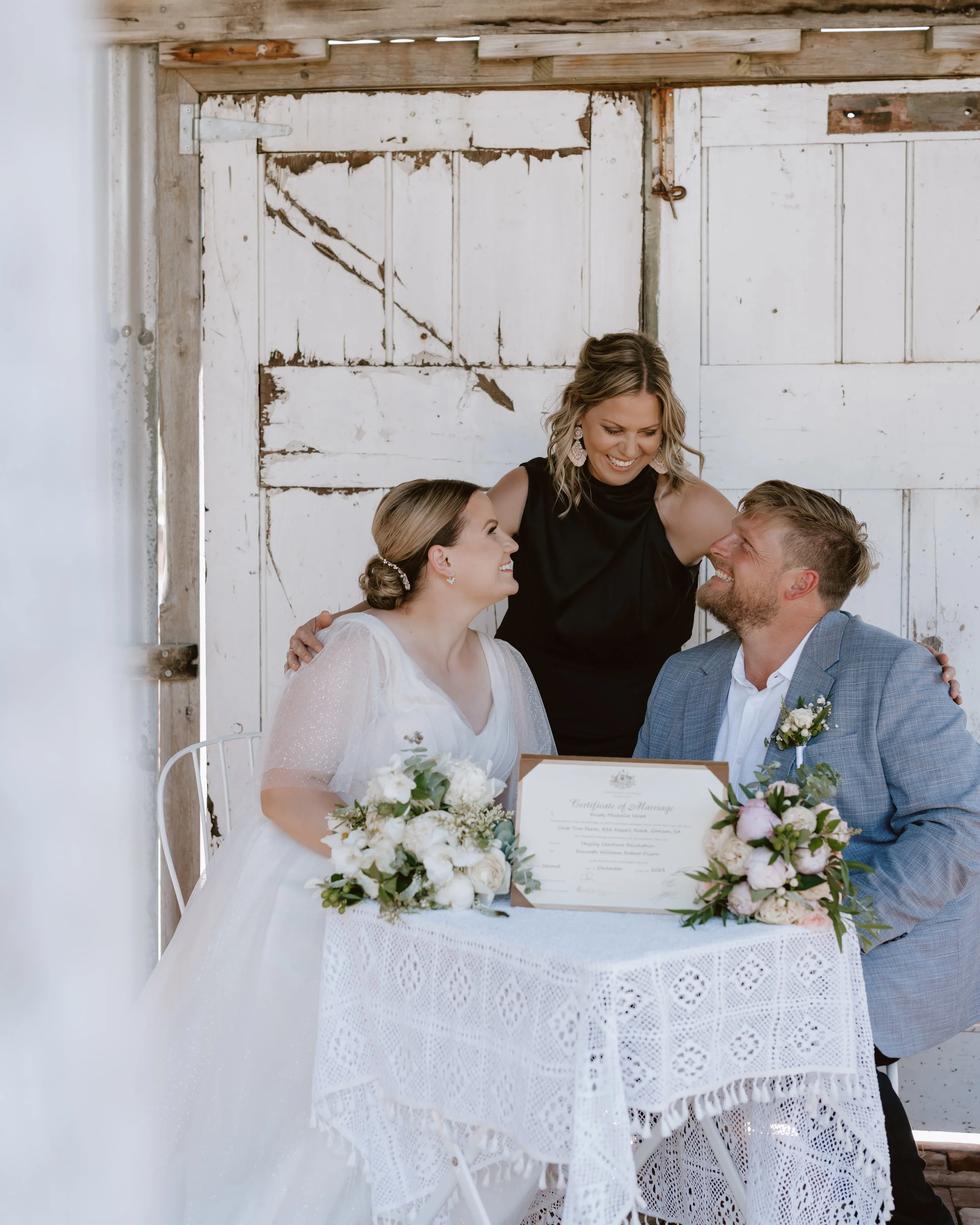Two women in white dresses sharing an intimate moment outdoors beneath green leafy vines, one kissing the other's forehead, both smiling affectionately.
