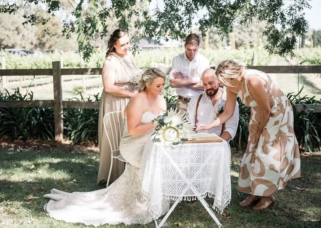 A couple sitting at a wedding table signing marriage legal papers with Kristy the Celebrant.
