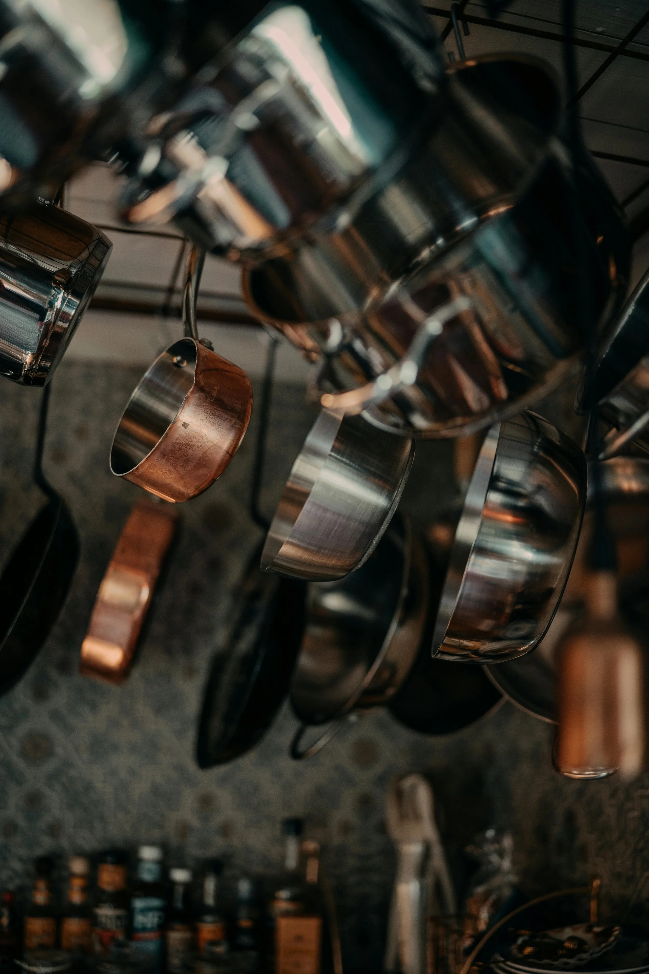 A collection of hanging metal mugs and cups, viewed at an angle, with a storage area and bottles in the background.