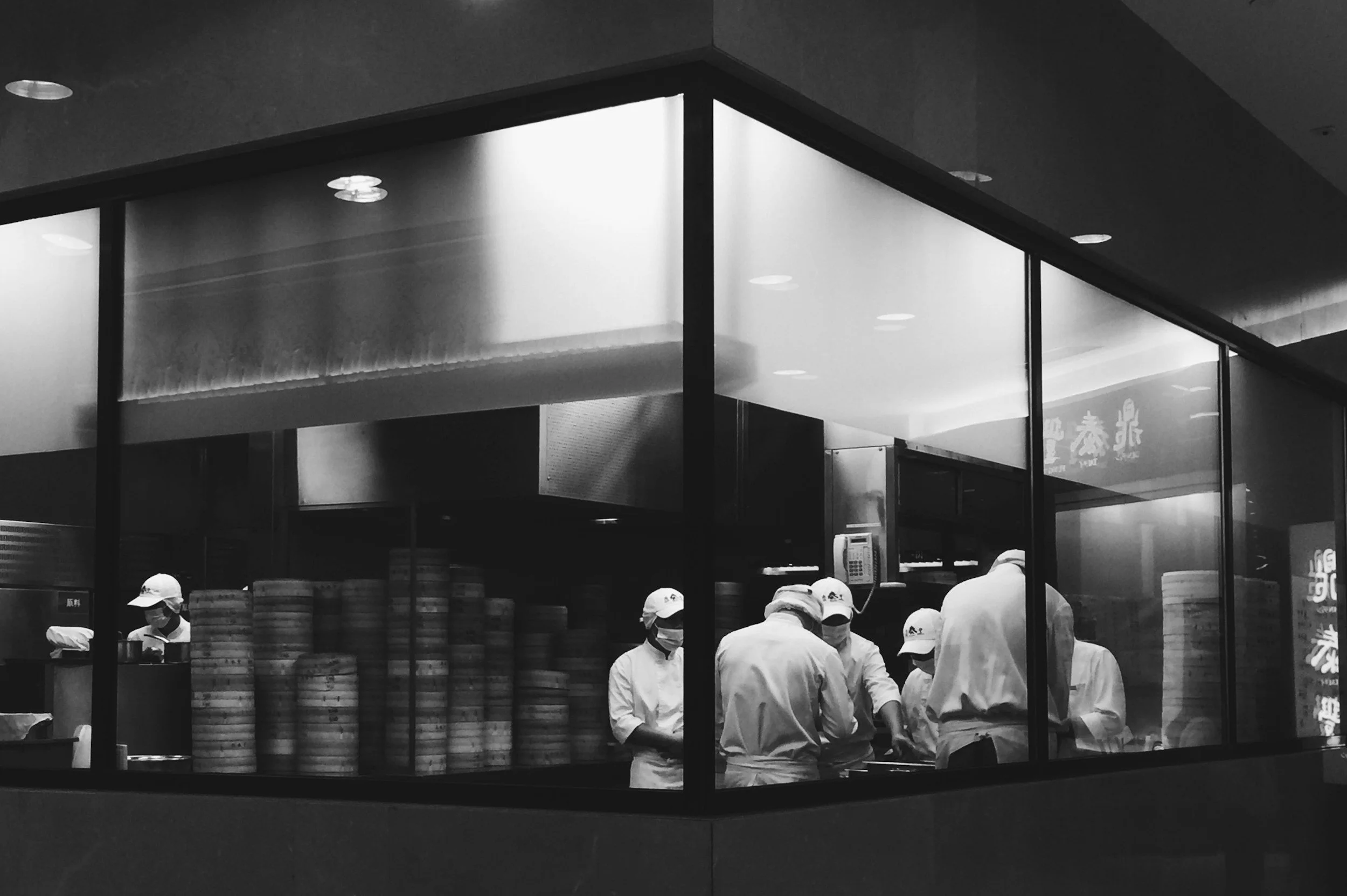 Black and white photo of a restaurant kitchen with chefs preparing food, stacks of bamboo steamers, and a glass wall showing the interior from outside.